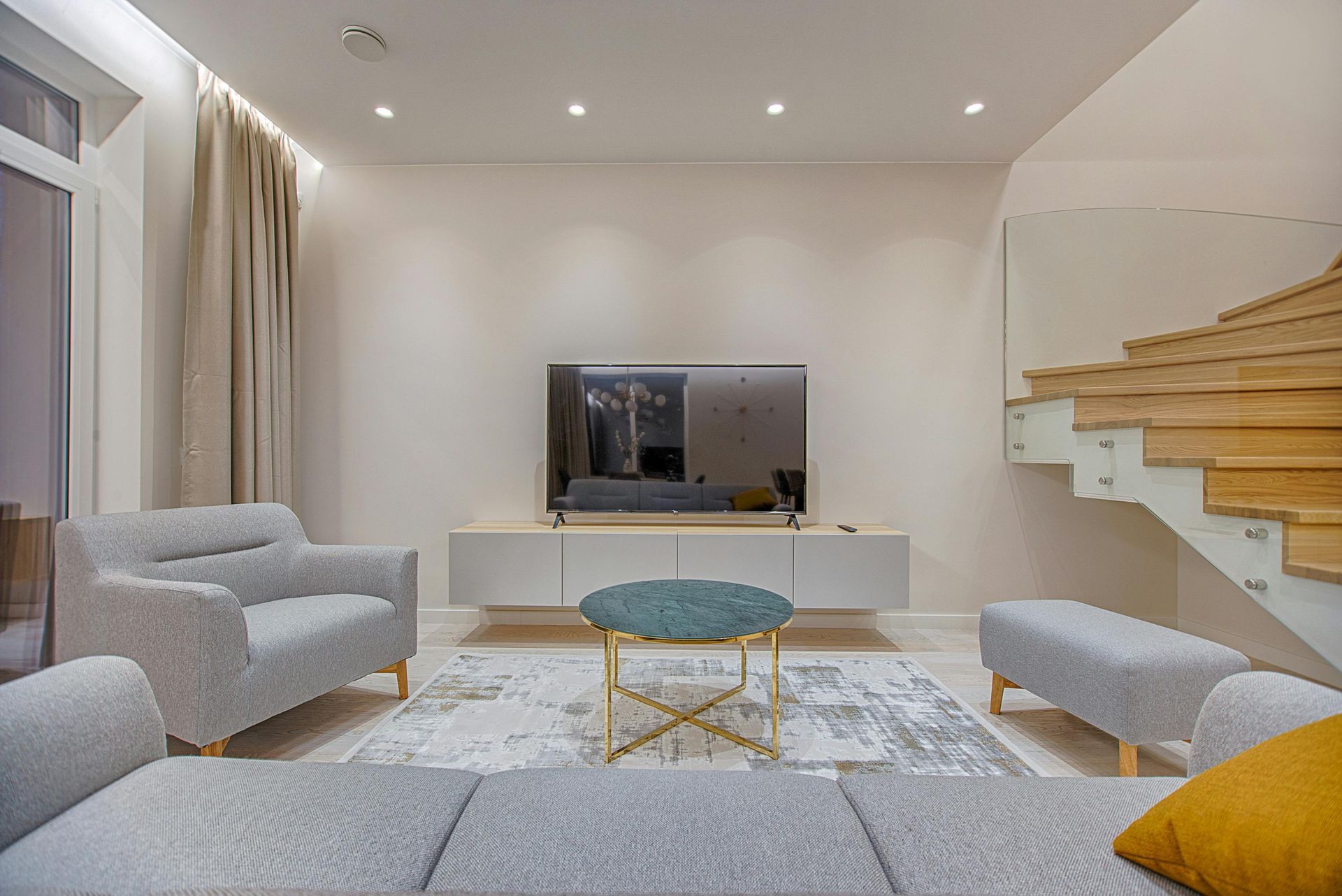 Modern living room with gray sofas, white walls, a TV console, and a small teal coffee table beneath a staircase.