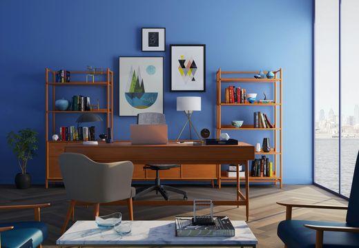 Modern home office with blue walls, wooden desk, bookshelves, framed art, and a city view through tall windows