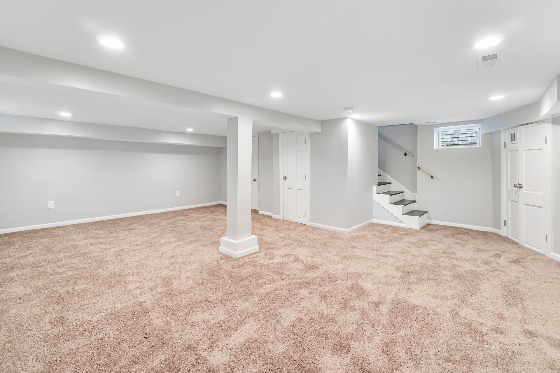 Empty finished basement with beige carpet, white walls, support columns, recessed lights, and stairs to an upper level