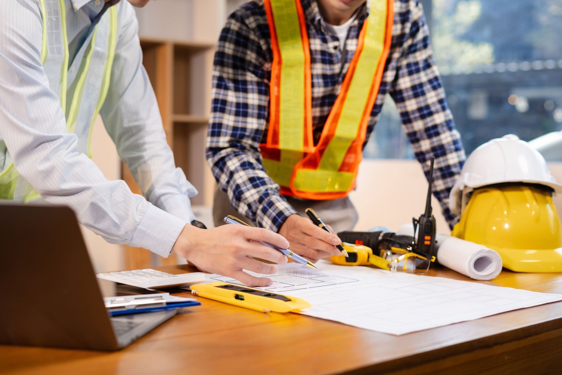 Construction workers reviewing plans at a table with tools, laptop, and hard hats on a job site