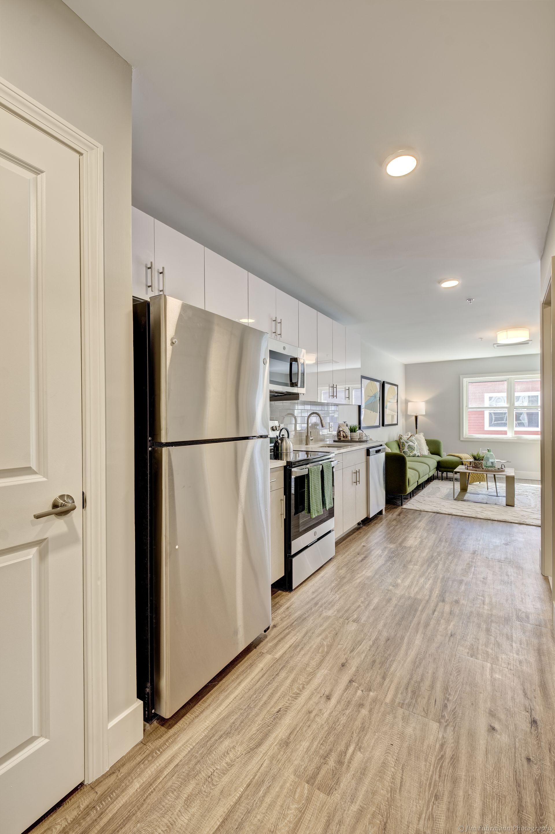 A small, modern kitchen with white cabinets and stainless steel appliances opens to a living room with a green sofa and a window.