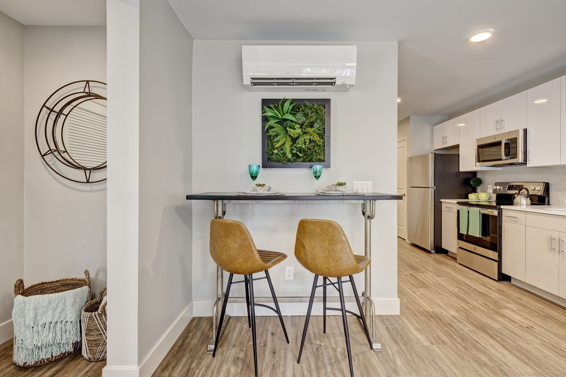 A modern dining area with a small black table, two brown chairs, a green plant wall, and a white kitchen in the background.