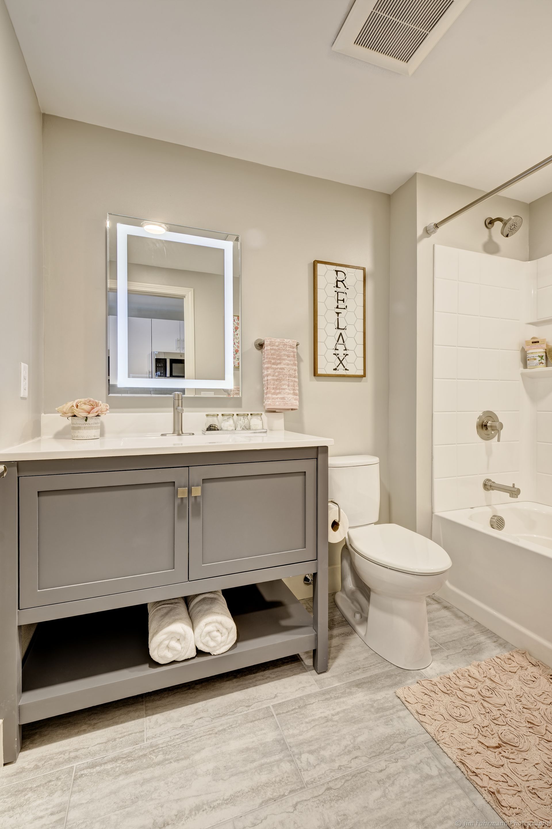 A modern bathroom with a gray vanity, white countertop, and backlit mirror. Includes a toilet, shower, and light gray flooring.