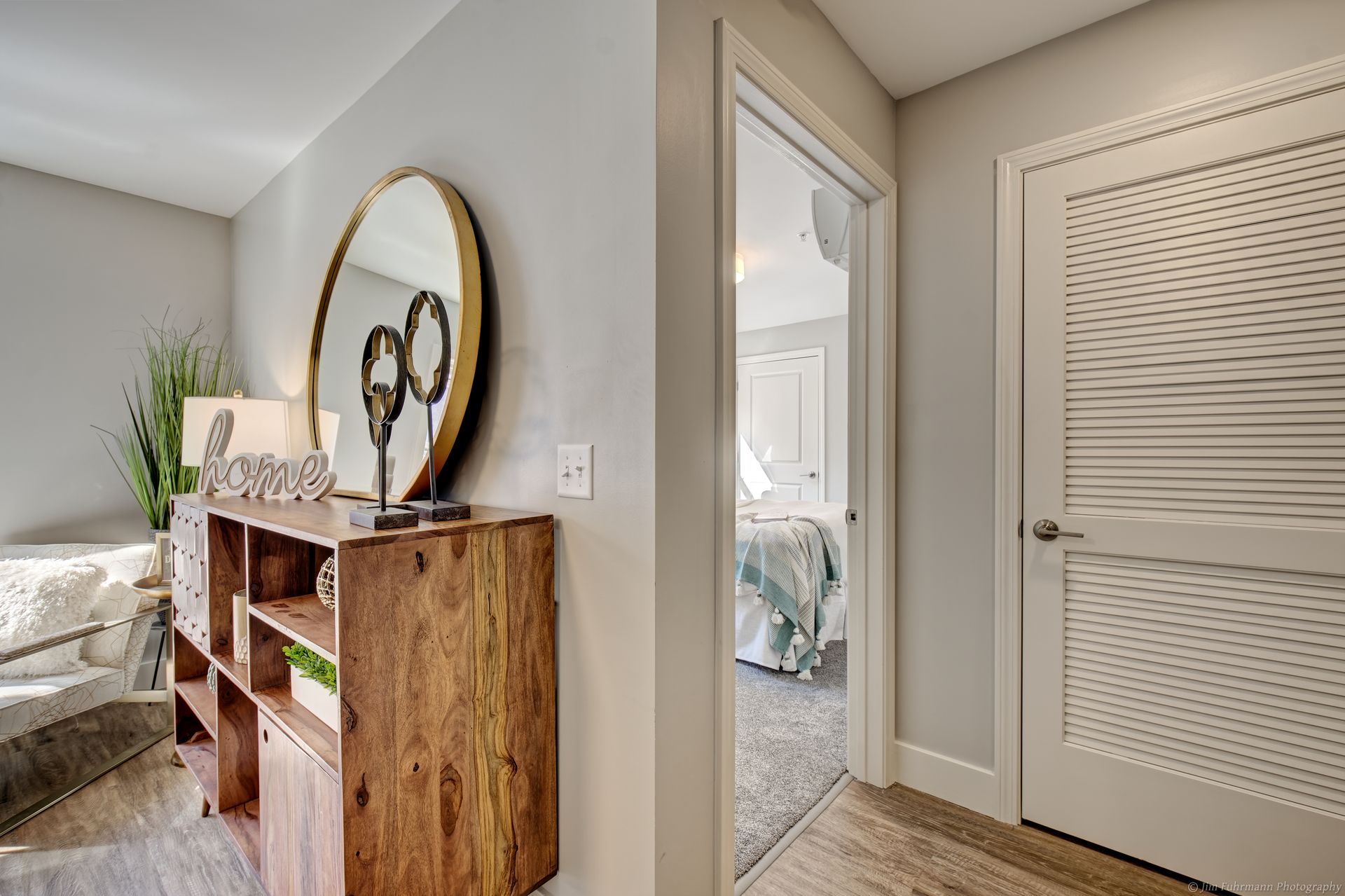 A hallway with a wooden cabinet, round mirror, and a view into a bedroom with a bed.