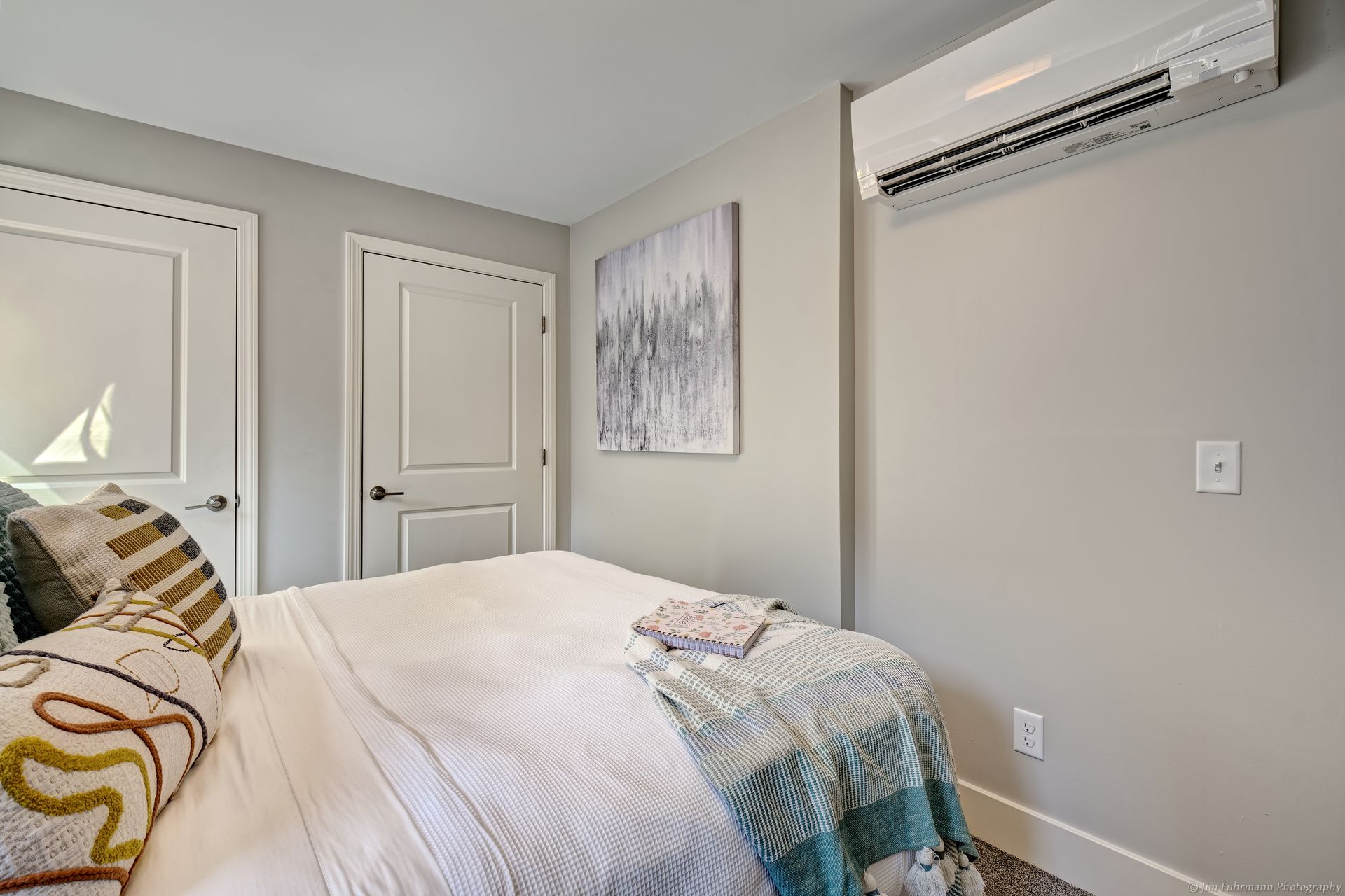 Bedroom with a bed, white doors, gray walls, and a wall-mounted air conditioner. A piece of art hangs above the bed.