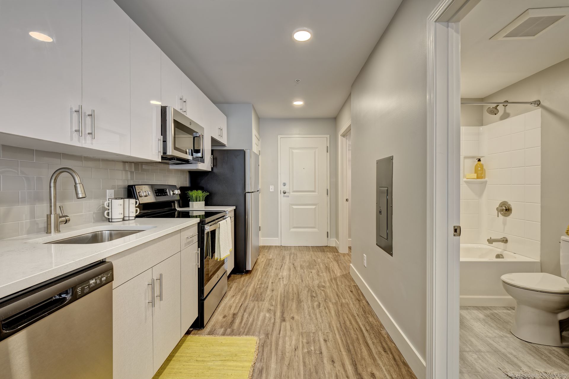 Small, modern apartment kitchen and bathroom. Features white cabinets, stainless steel appliances, and light gray walls with wood-look flooring. The bathroom is visible on the right.