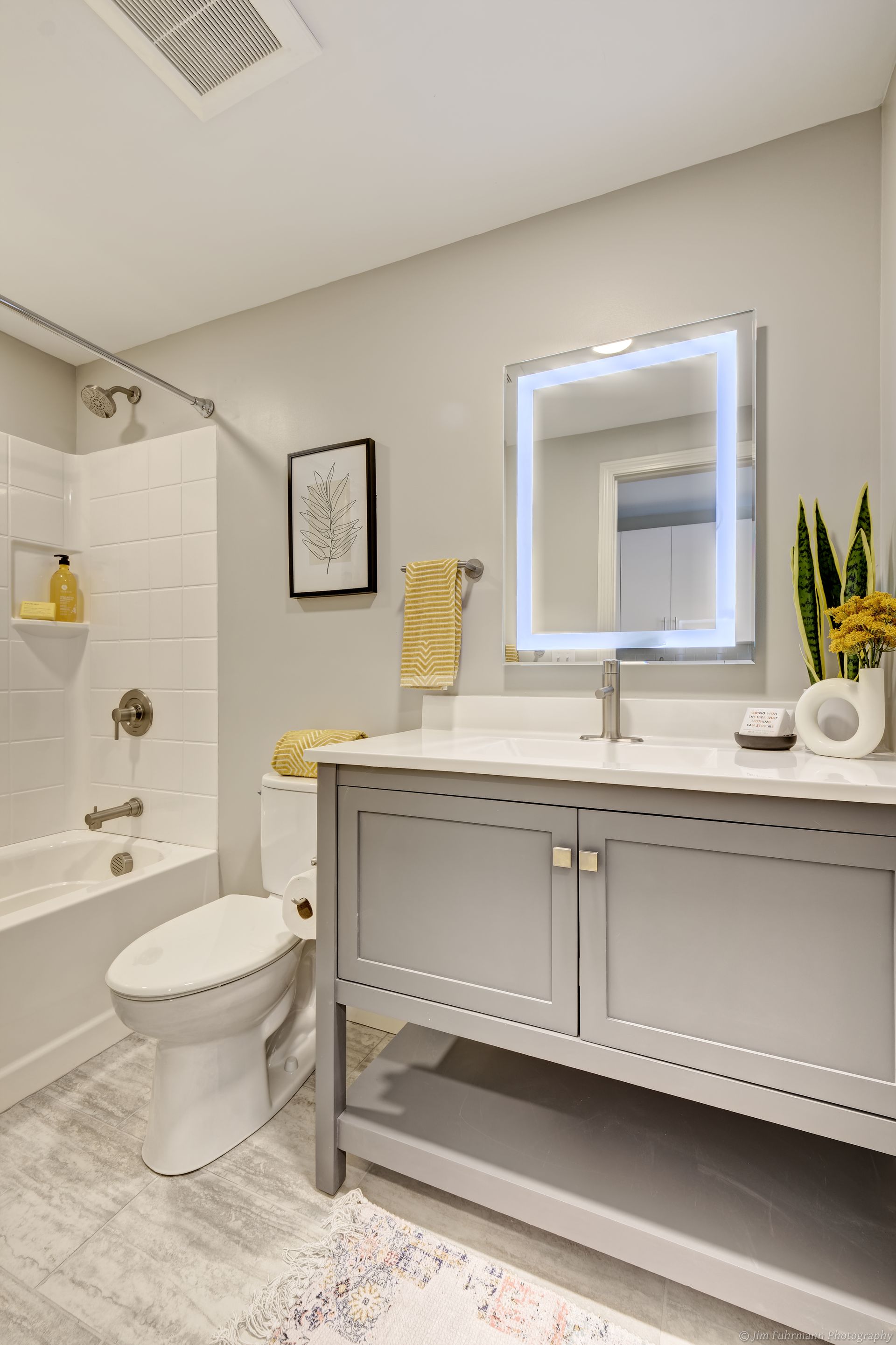 A modern, bright bathroom with a gray vanity, backlit mirror, white toilet, and subway tile shower. Yellow accents pop against the neutral color palette.