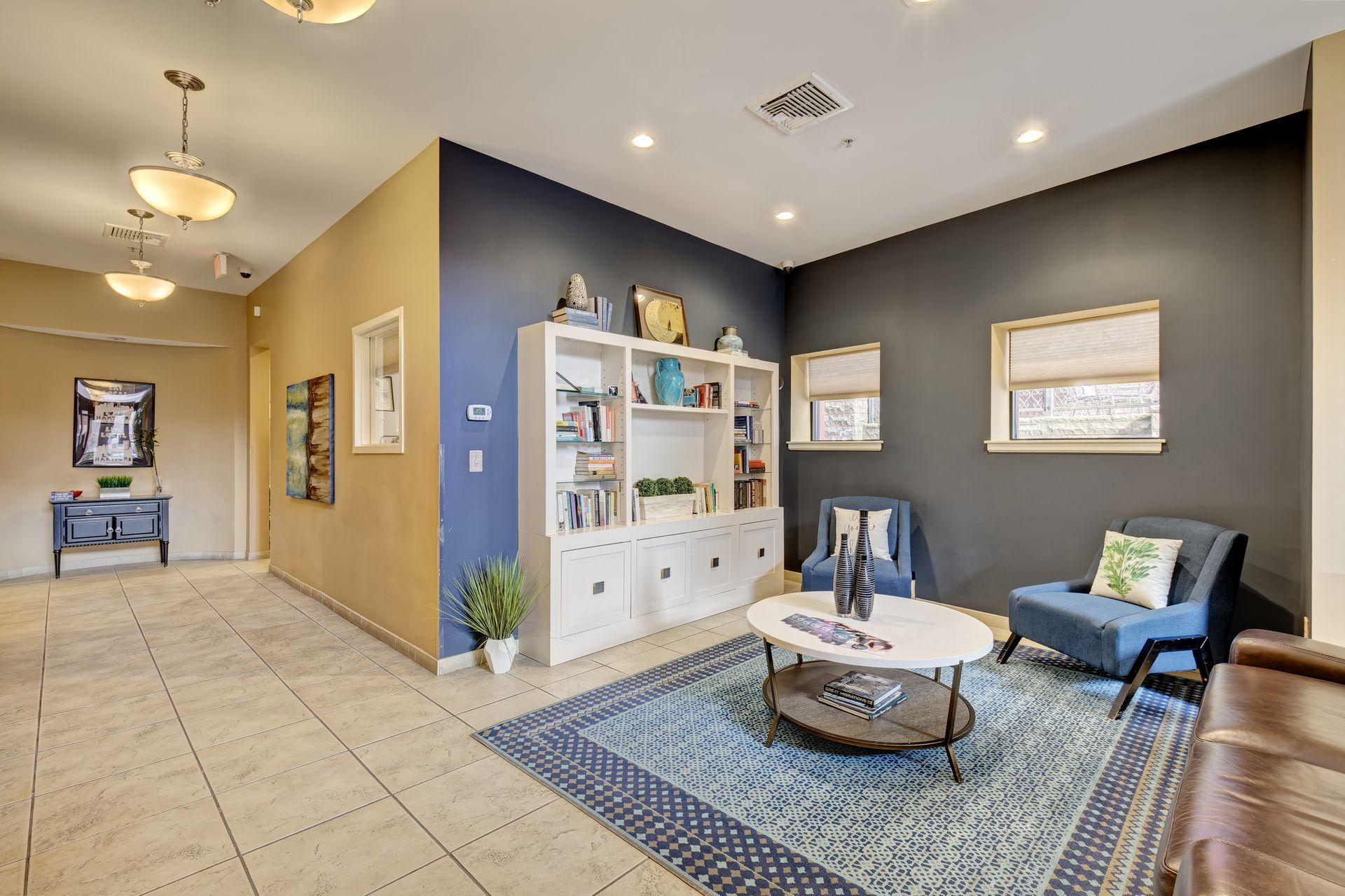 A well-lit waiting area with blue and tan walls, a white bookshelf, and blue seating arranged around a coffee table on a patterned rug.