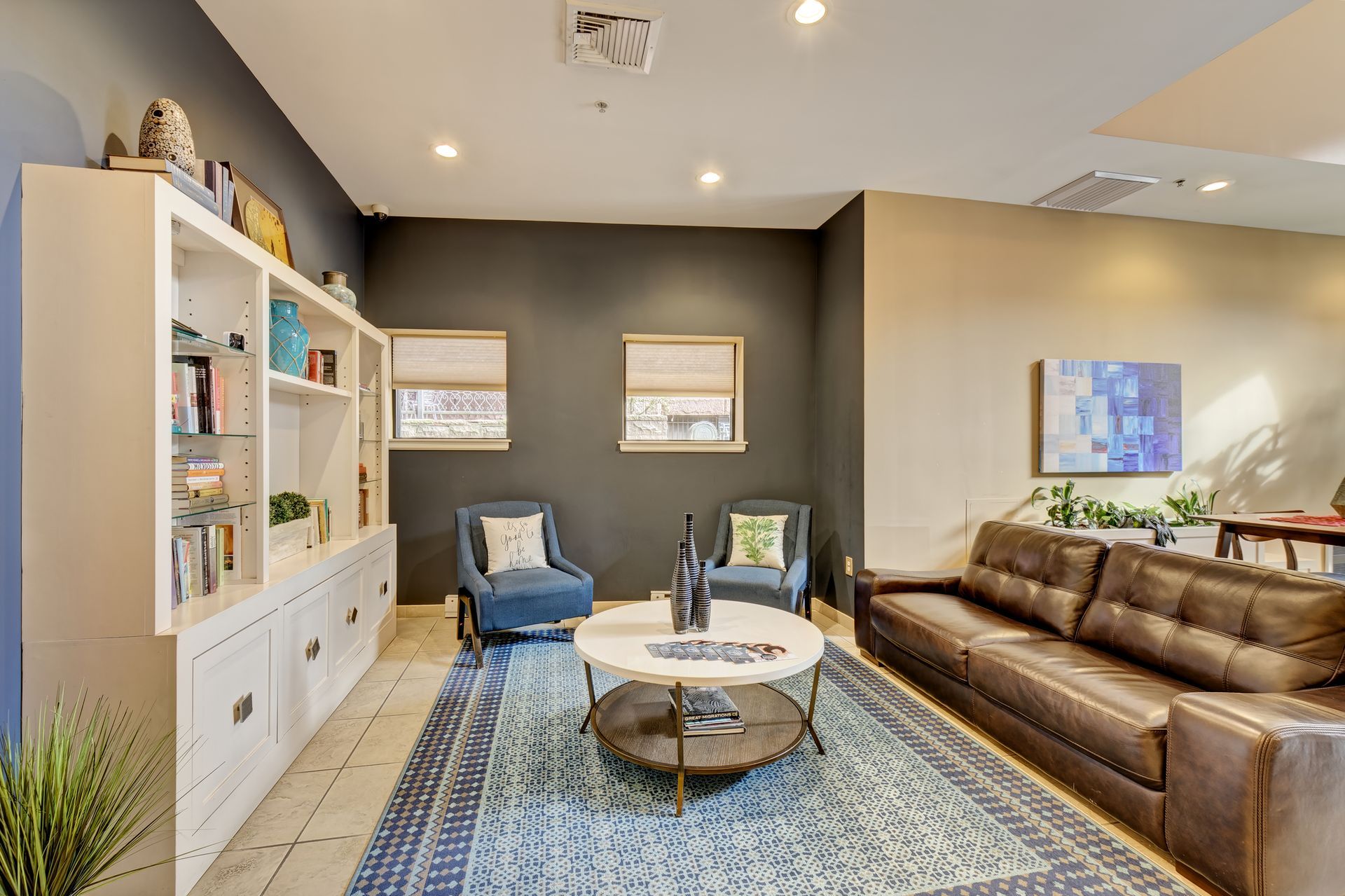 A waiting area with blue and brown decor. Features a leather couch, blue armchairs, a coffee table, and a bookshelf.