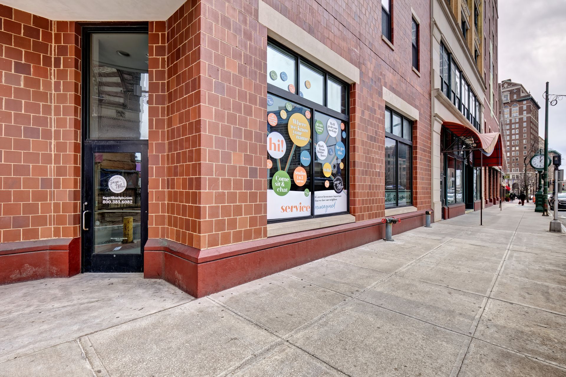 Exterior of a brick building with a glass door and windows. The windows display colorful decorations. A sidewalk runs in front of the building.