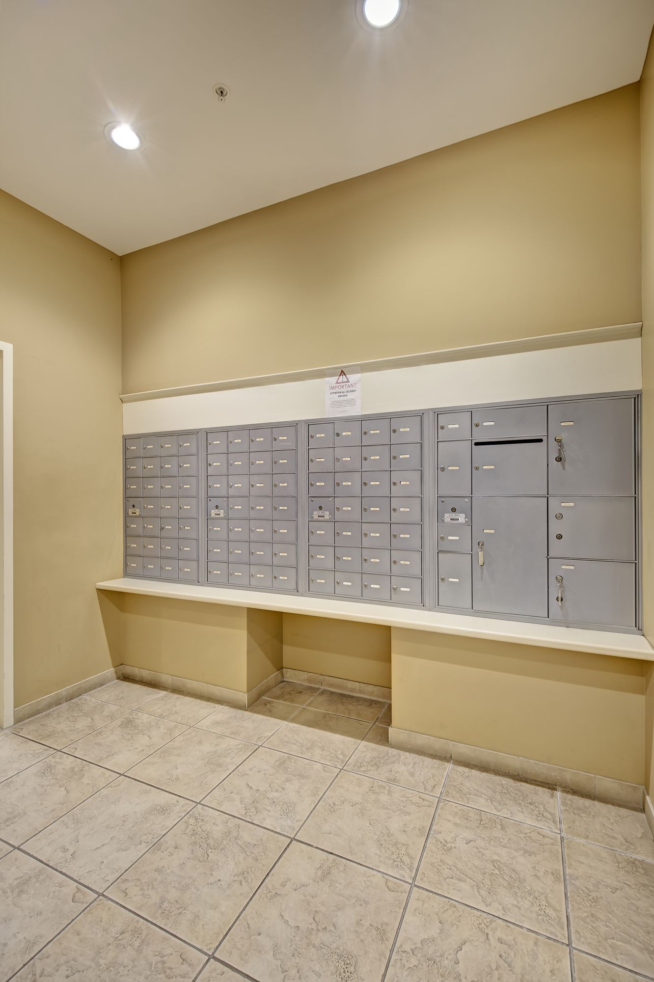 Mailboxes in a hallway, featuring a row of metal boxes with a countertop below. Beige walls and tiled floor.