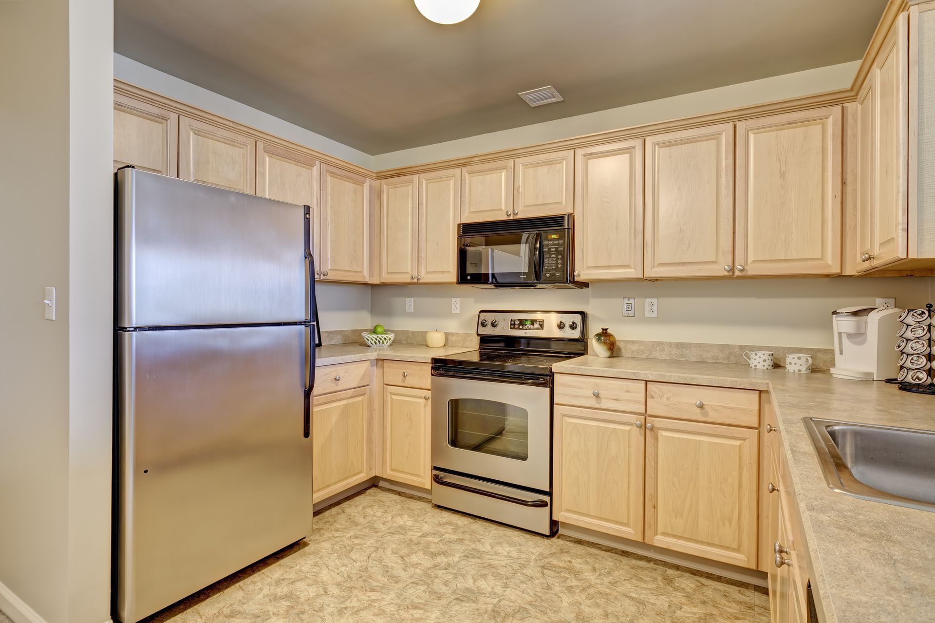 Stainless steel appliances in a light-wood toned kitchen. Cabinets, counters, and a refrigerator are visible.