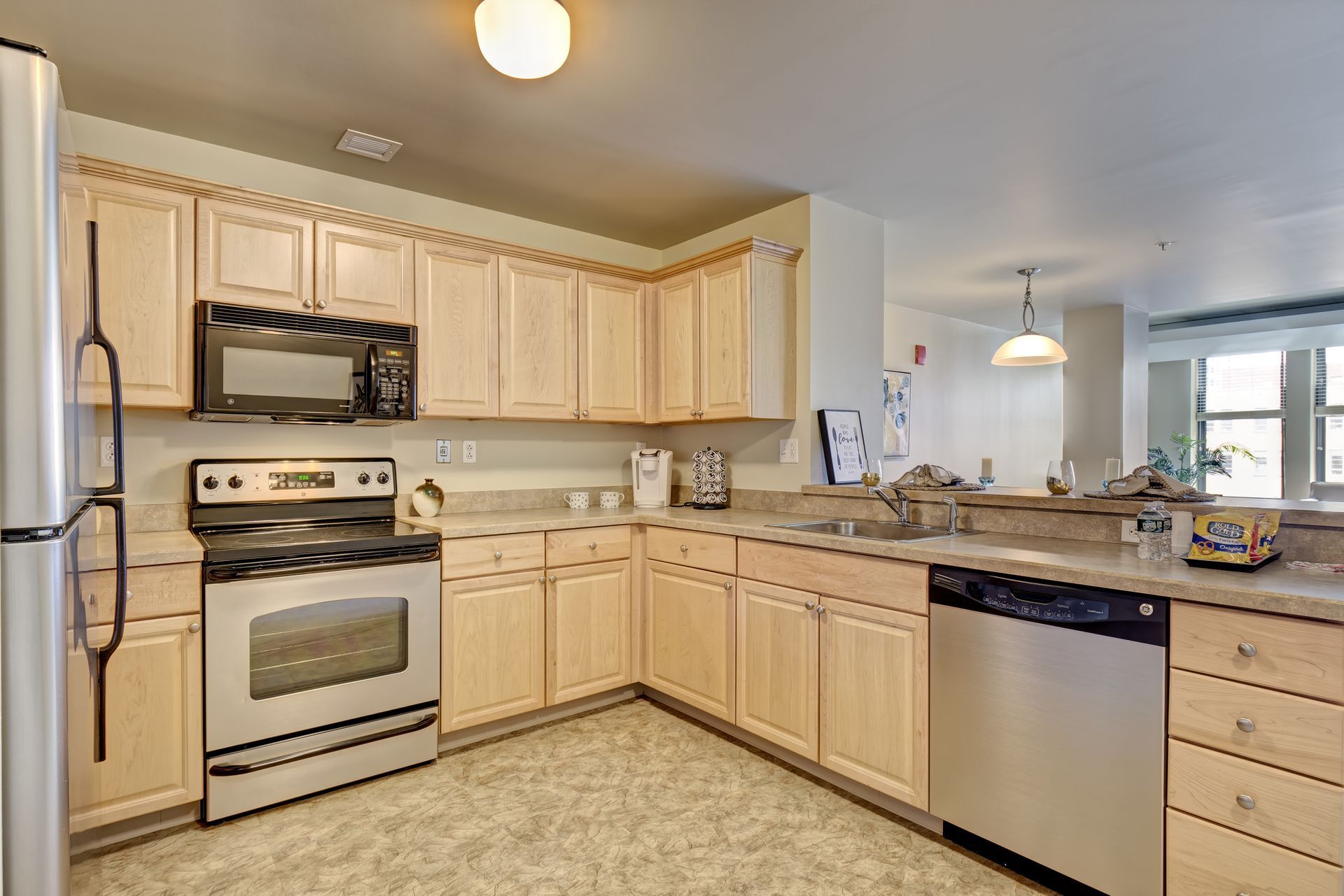 A kitchen with light wood cabinets, stainless steel appliances, and a neutral countertop. The room opens to a dining area.