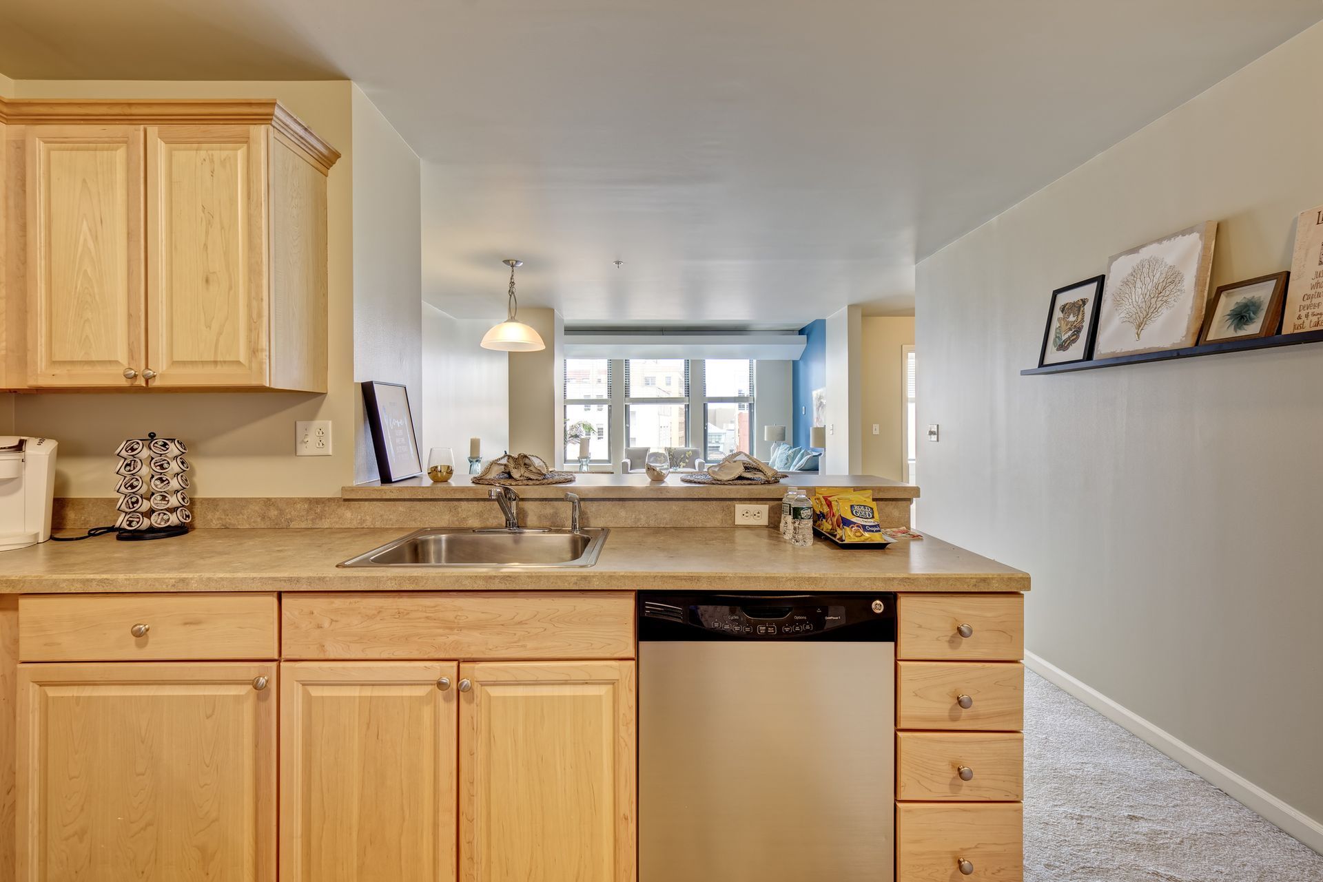 Kitchen with light-colored wooden cabinets, a sink, and a stainless steel dishwasher. A view through an opening leads to a dining area.