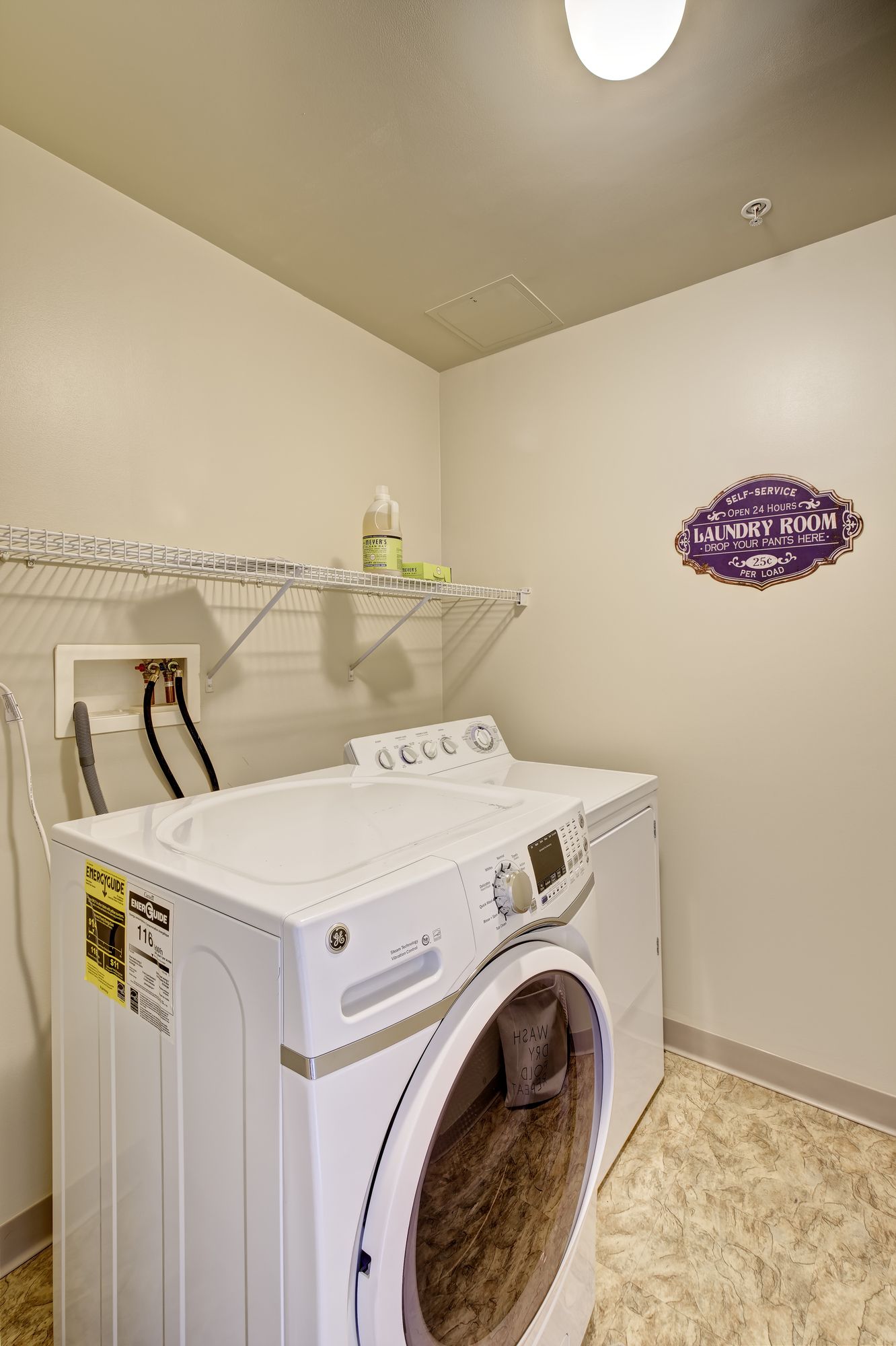 Laundry room with a white front-loading washing machine, wire shelving, and a decorative purple sign. The floor is light-colored with a speckled pattern.