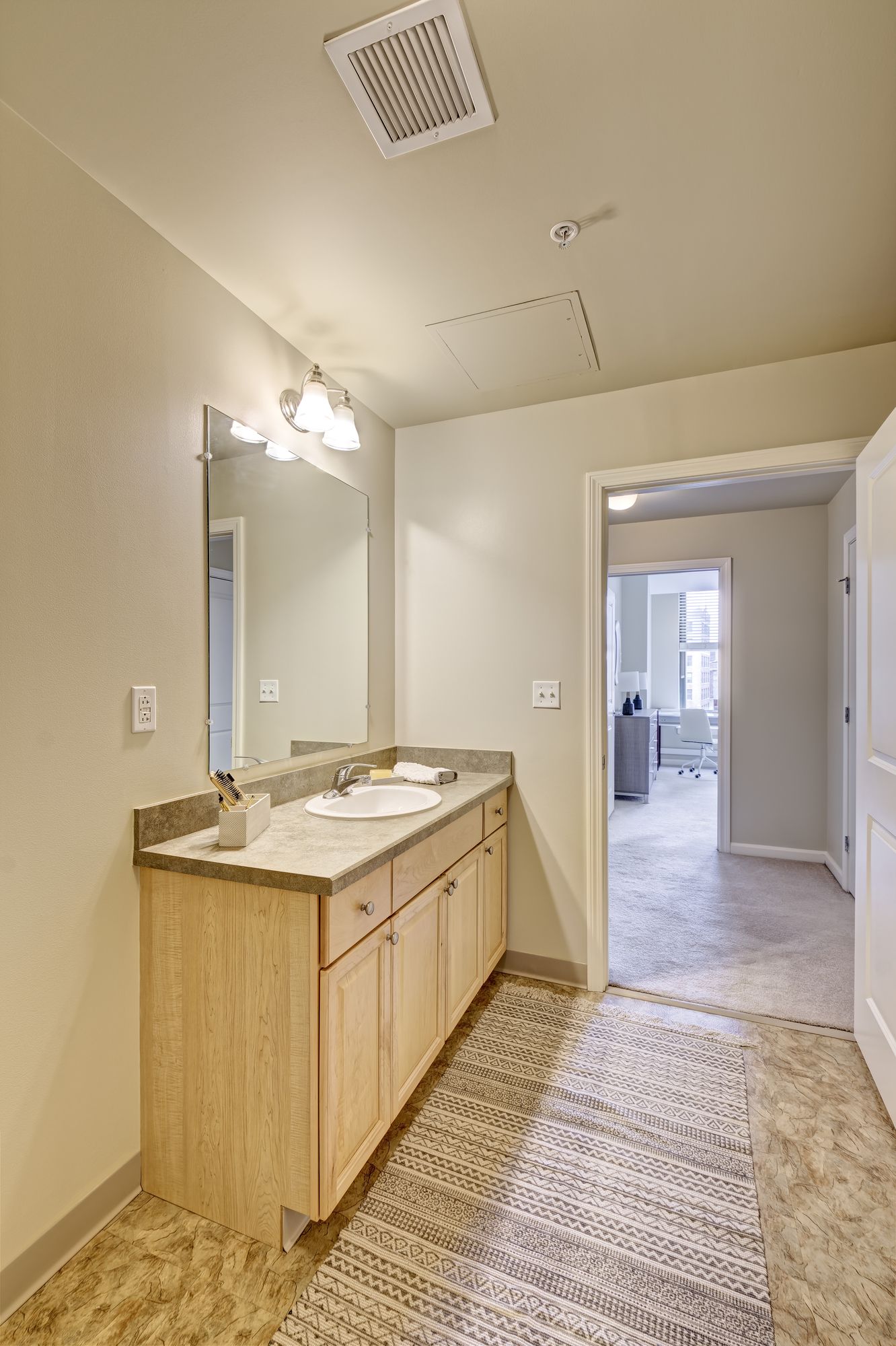 Bathroom with light wood vanity, long mirror, and patterned floor mat. A hallway is visible to the right.