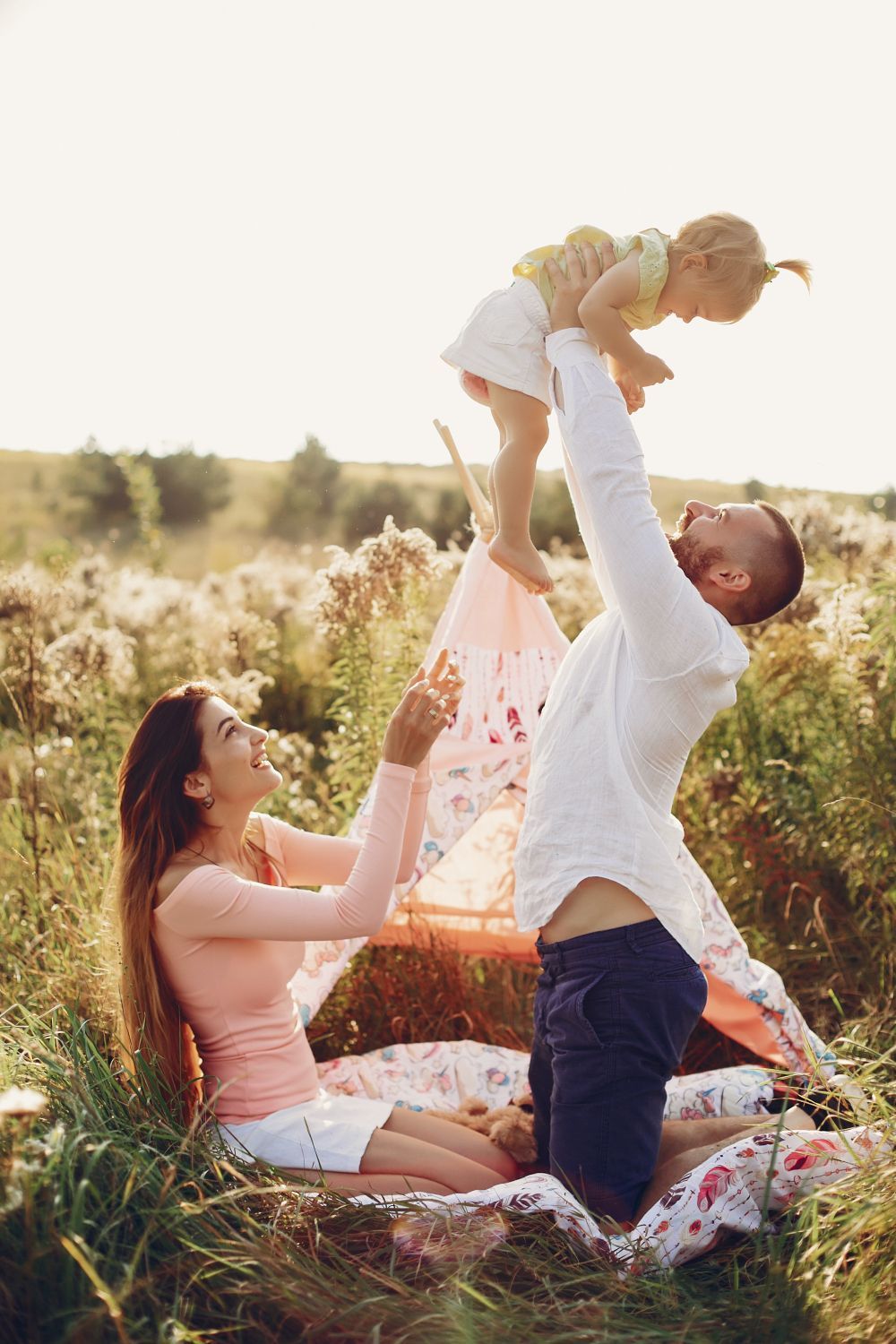 Family of three outdoors, playing in a sunny field. Man lifts a toddler, as woman watches joyfully from a blanket near a teepee.