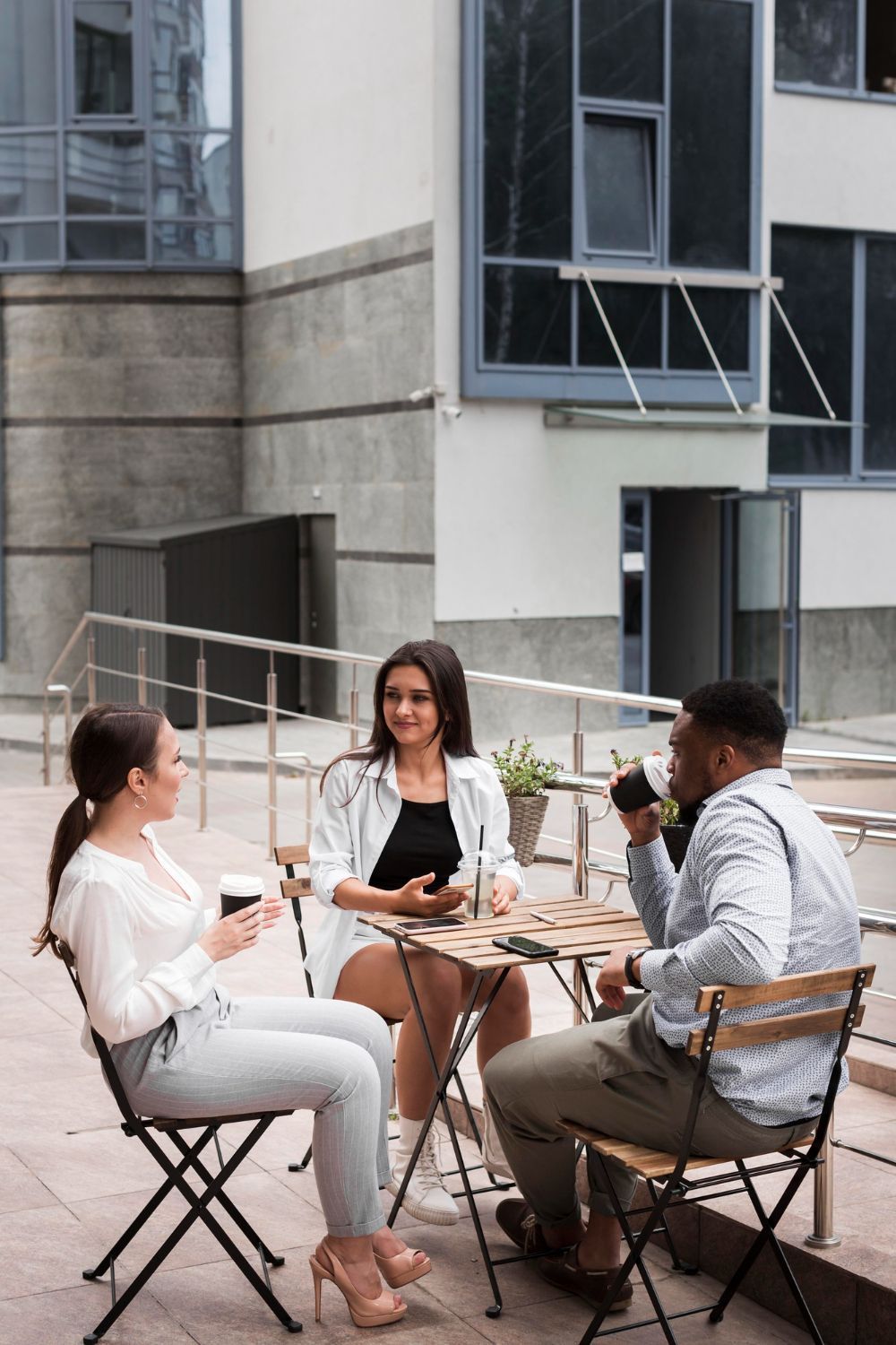 Three colleagues sitting at a small outdoor table, having a conversation. They are dressed in business casual attire, with the building in the background.