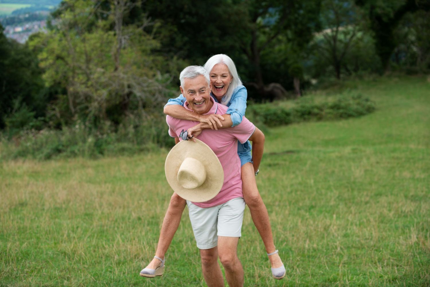 Elderly couple playfully enjoying the outdoors; woman riding on man's back in a grassy field, man wearing a hat.
