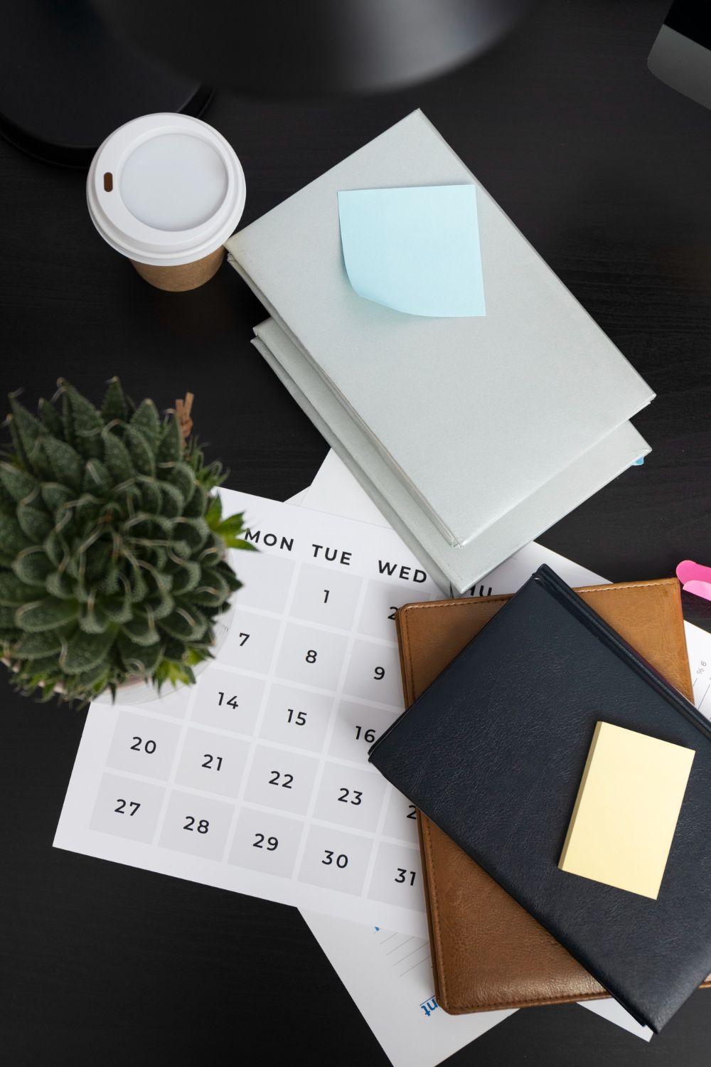 Desk scene with a succulent, coffee cup, stacked notebooks, calendar, and sticky notes.