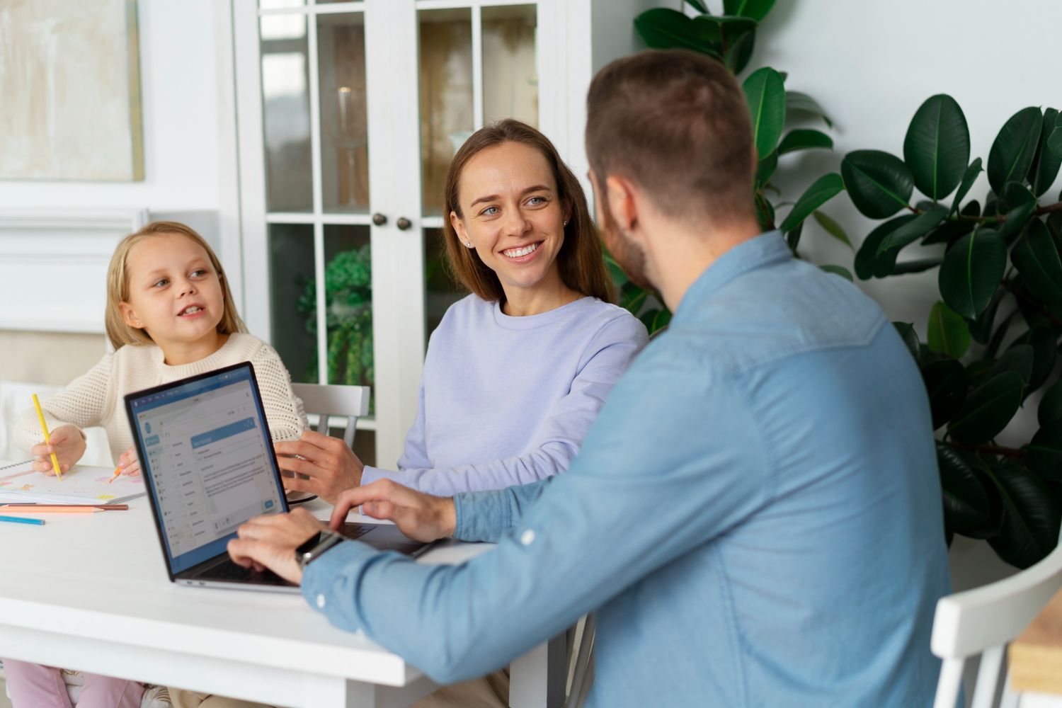 A family sits at a table: a father uses a laptop, a mother smiles, and a child draws. Natural light fills the room.