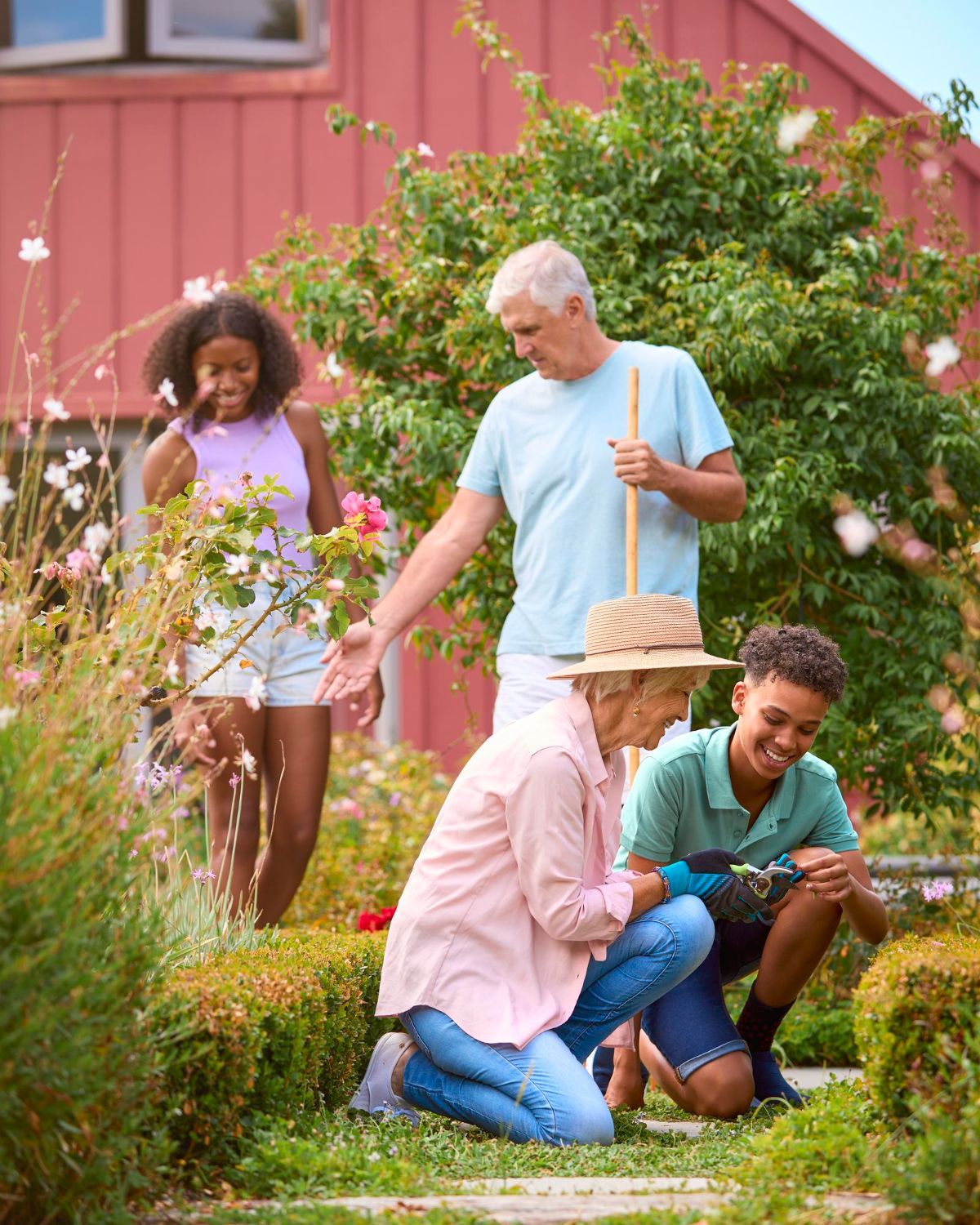 Family gardening together: grandparents and grandchildren in a garden. One grandmother kneels, using a tool; others watch and smile.