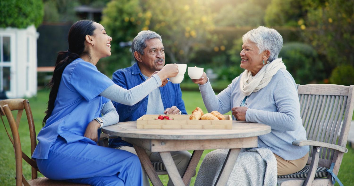 A caregiver, older man, and woman toast with mugs at an outdoor table. They are smiling in a sunny garden setting.