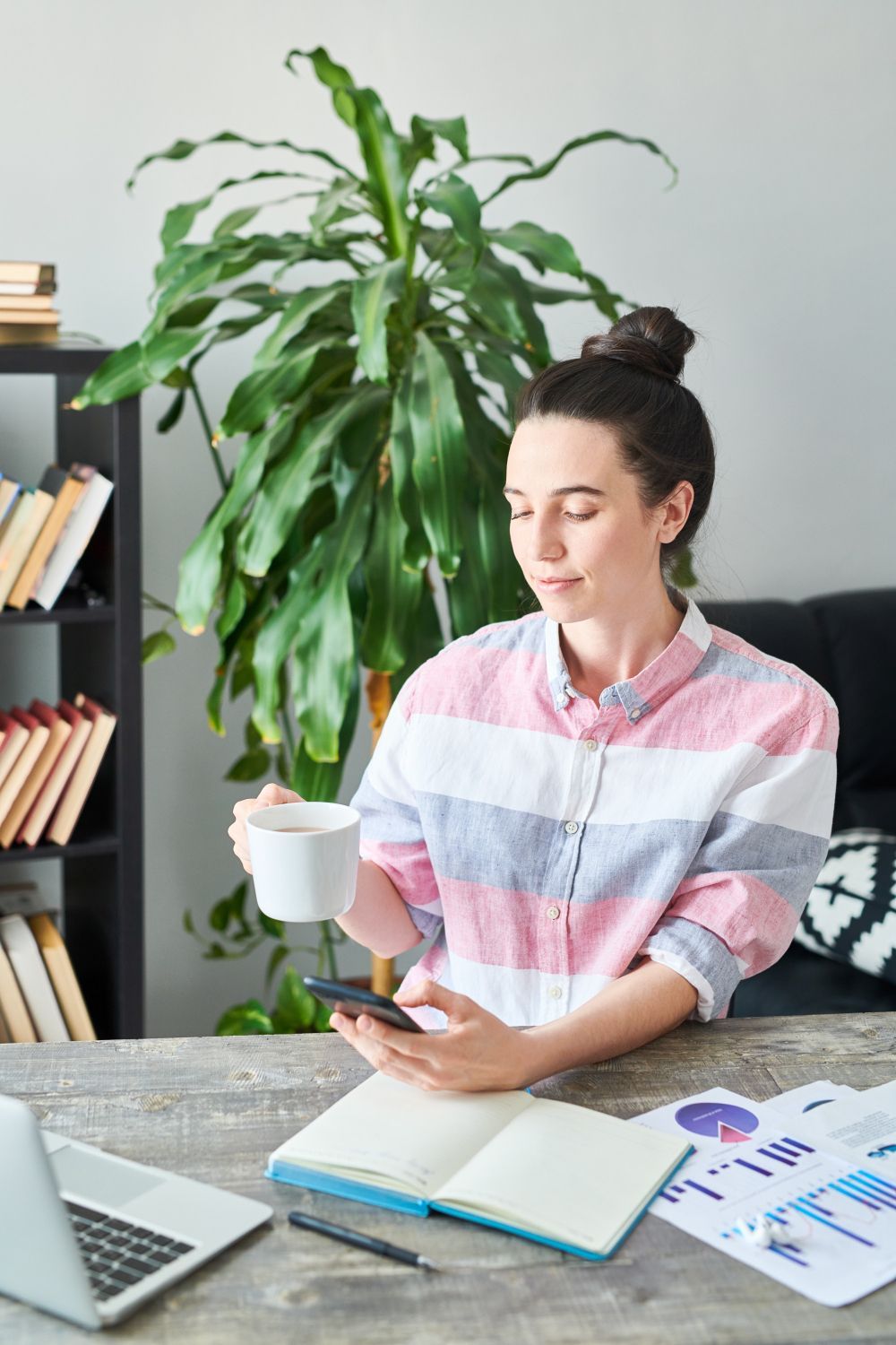 Woman in striped shirt holding coffee and phone at a desk, looking at the phone. Notebook, laptop, and charts on desk. Indoor setting.