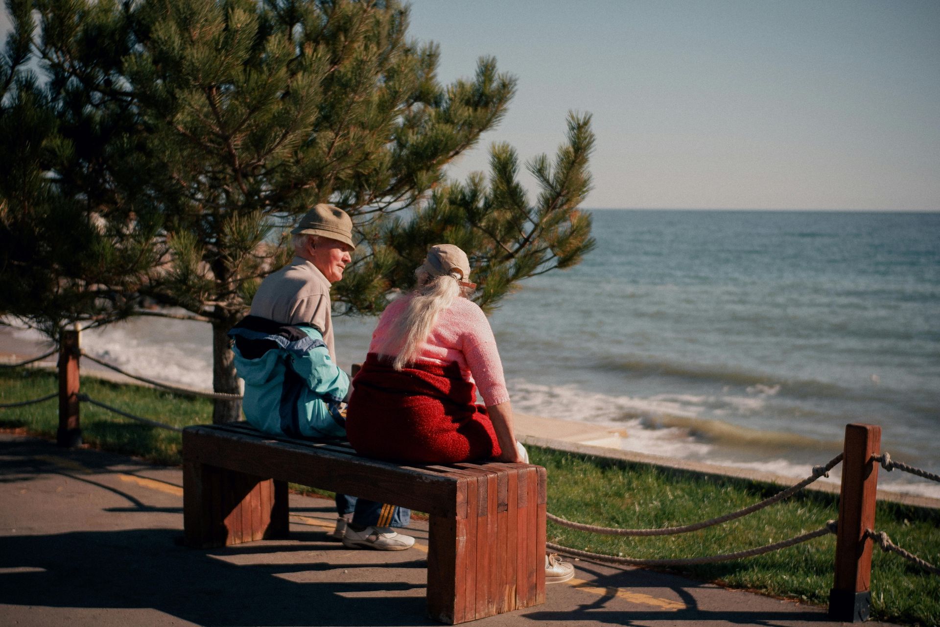 Elderly couple sits on a bench overlooking the ocean. One wears a fedora and blue jacket; the other, a pink shirt and red pants.