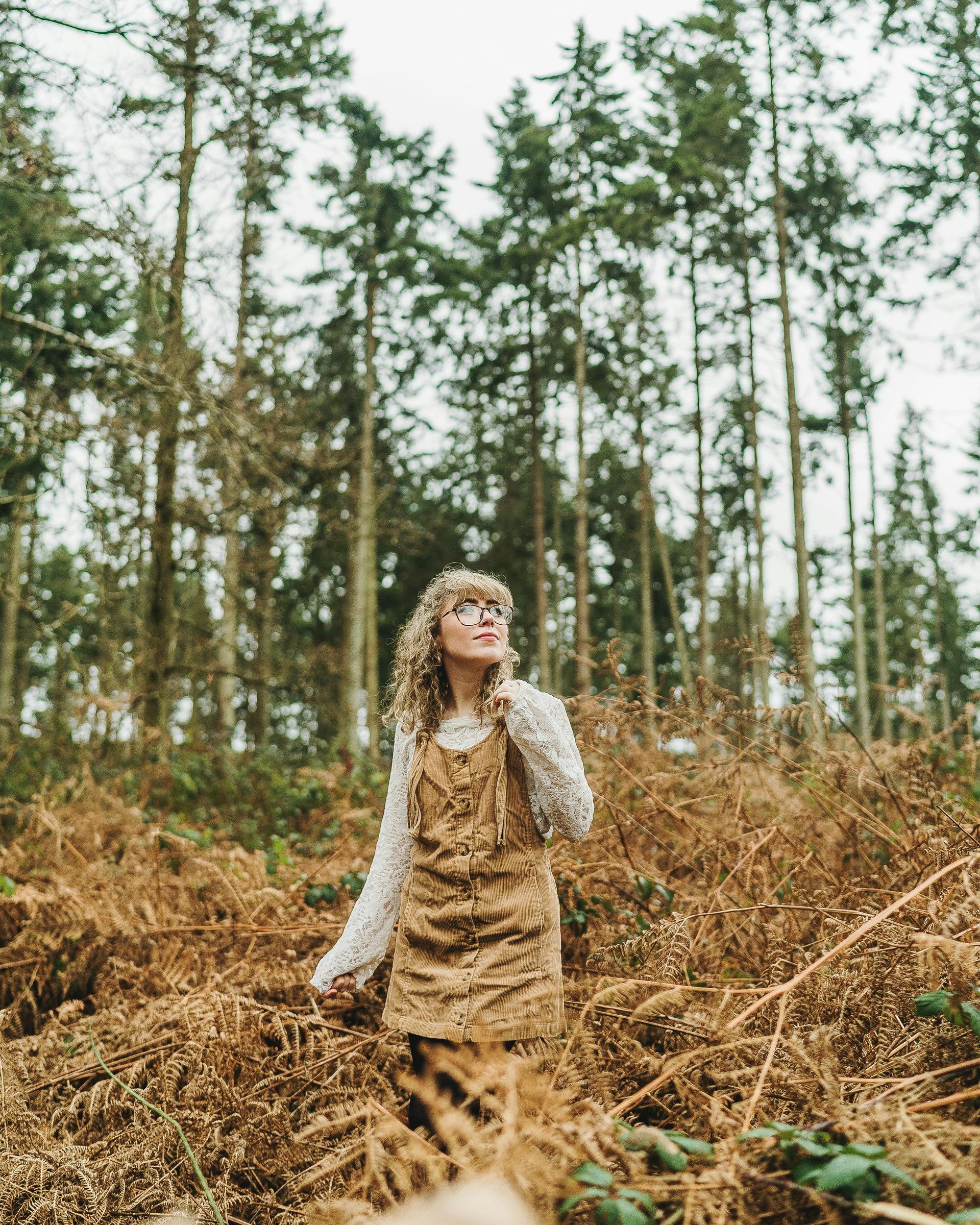 Woman with curly hair in a forest looks upward, wearing a white sweater under a brown corduroy dress.