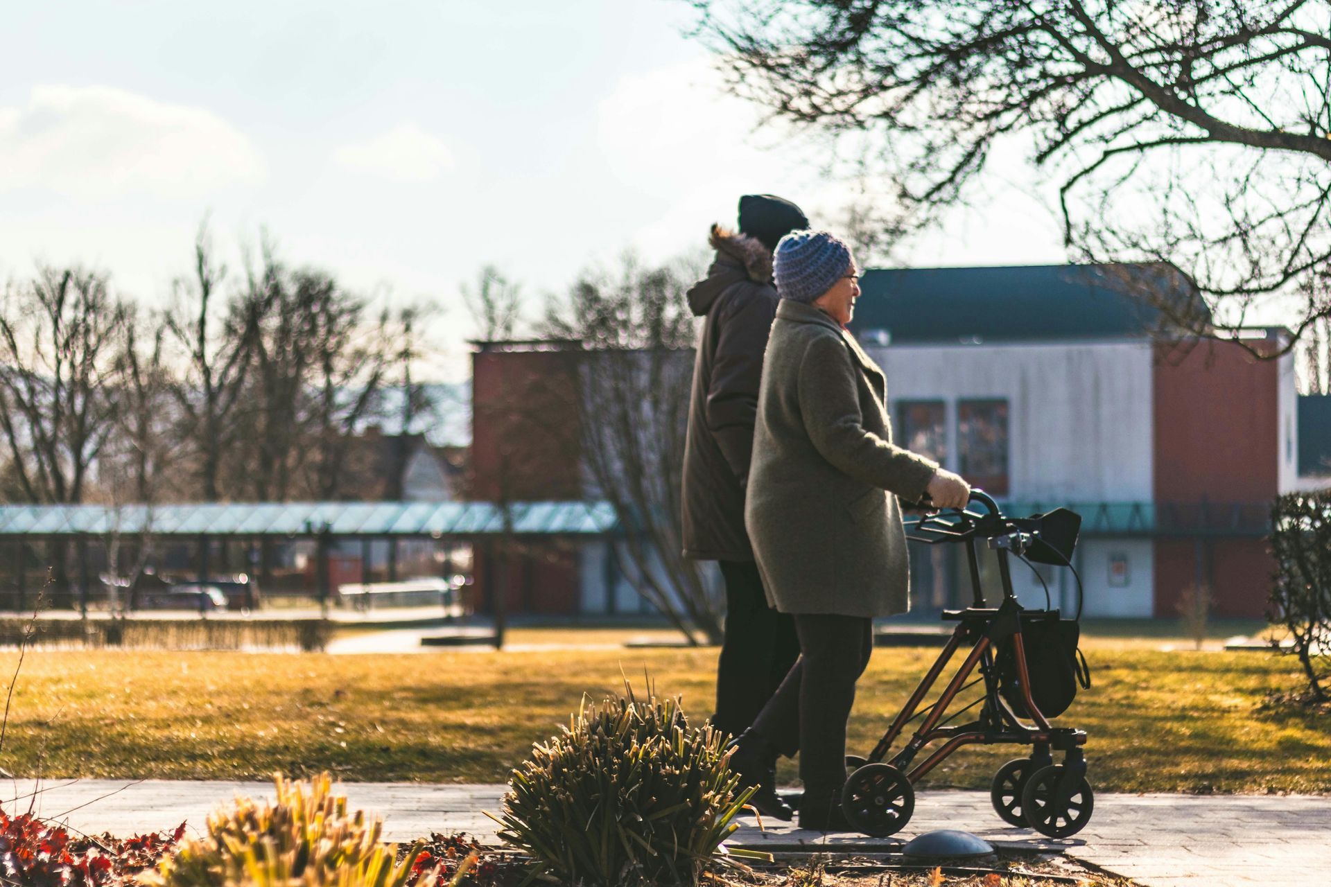 Two older adults walking outdoors, one using a walker. The setting is a sunny, grassy area with buildings in the background.