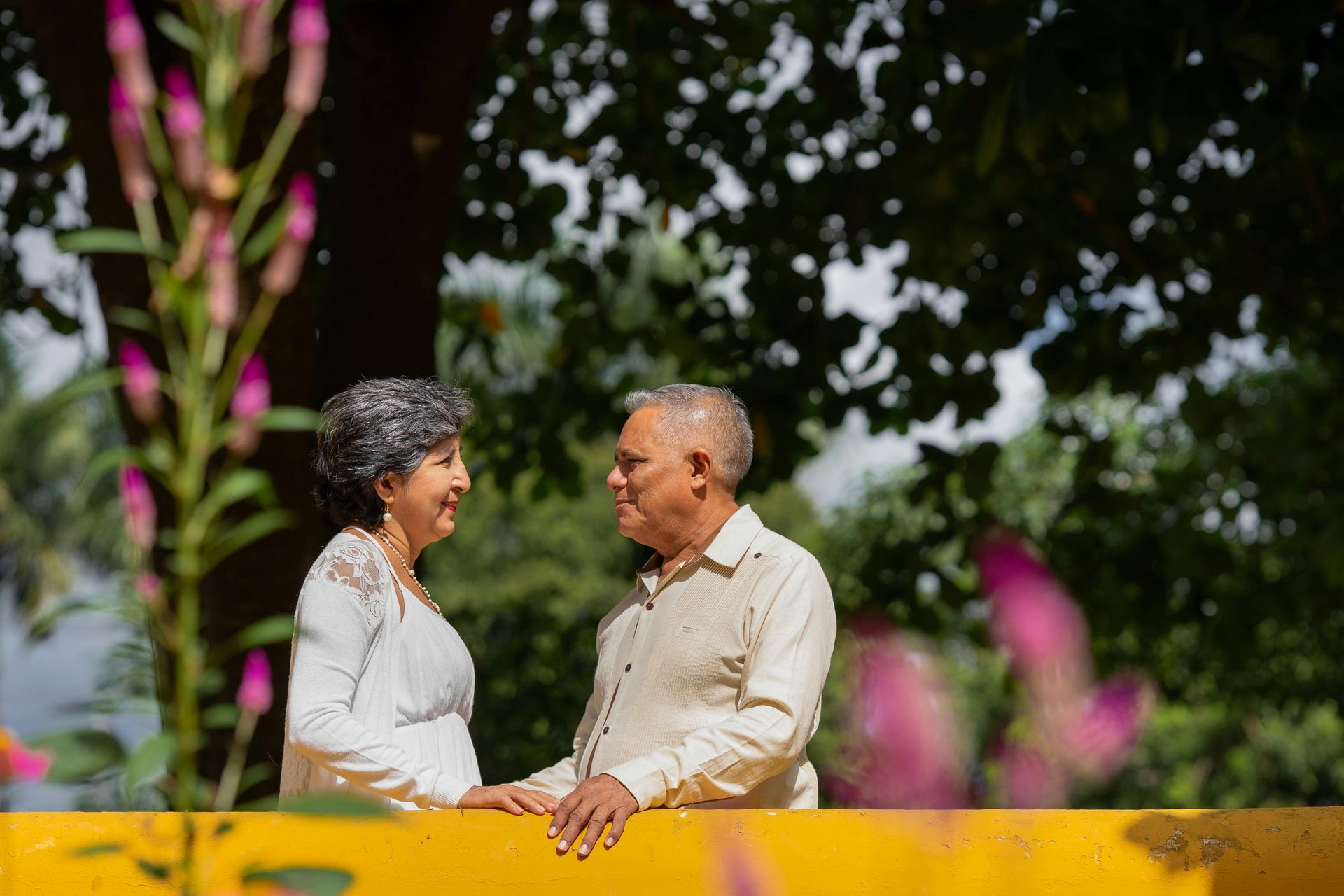 An elderly couple smiles at each other while standing near a yellow structure. Pink and purple flowers are in the foreground.