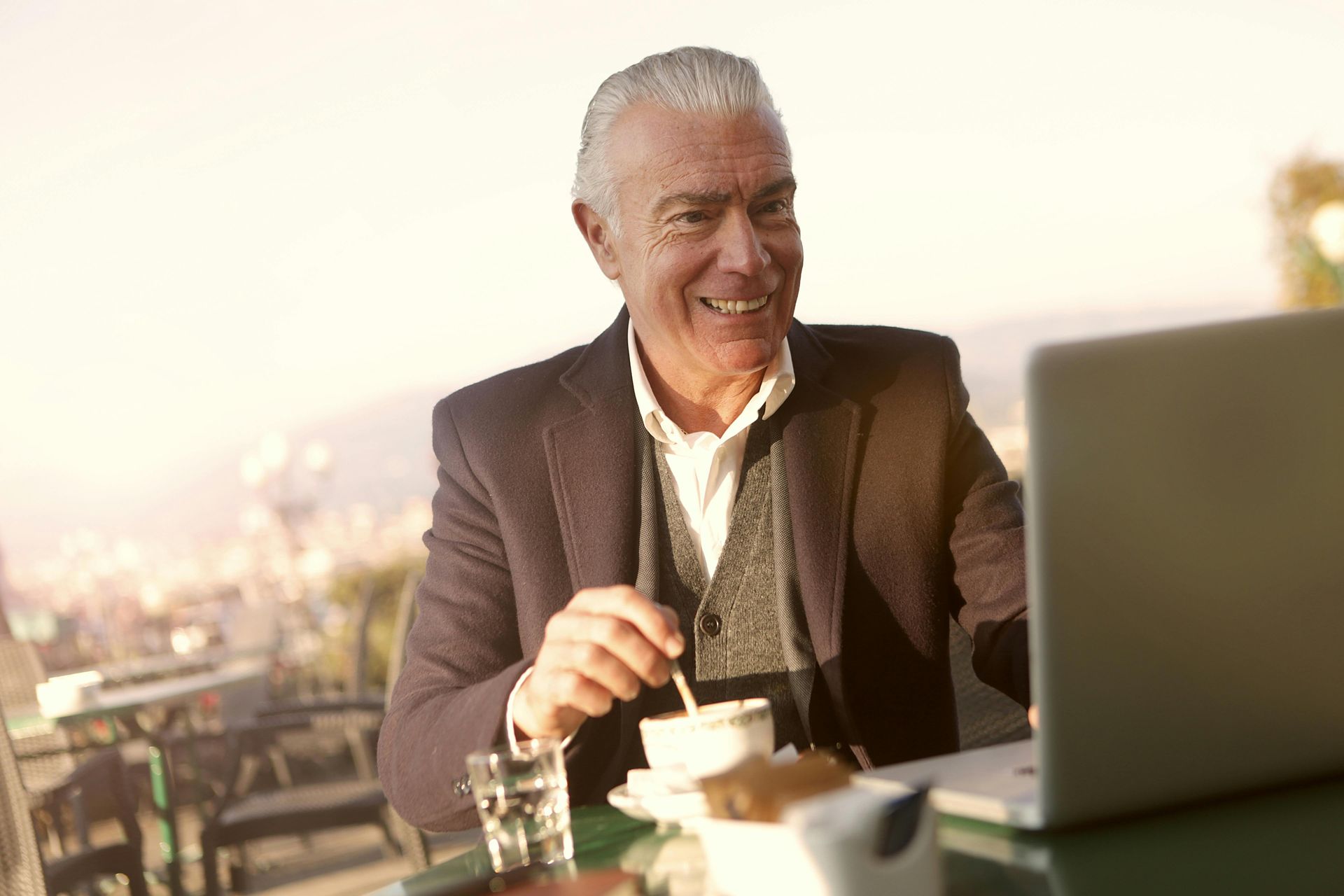 Smiling older man in a jacket stirs coffee while using a laptop at an outdoor cafe with a city view.