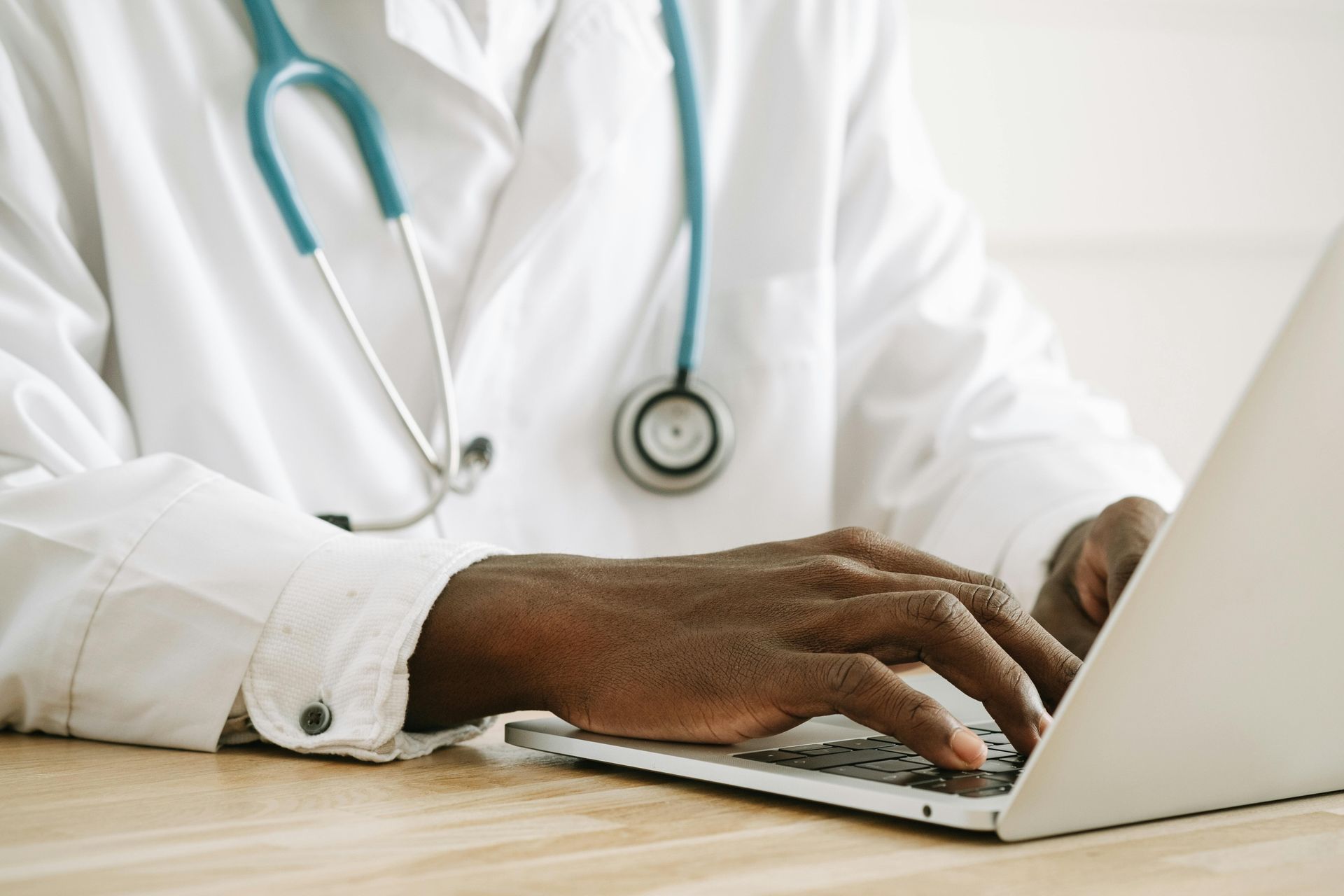 Doctor typing on a laptop at a desk, wearing a white coat and teal stethoscope.