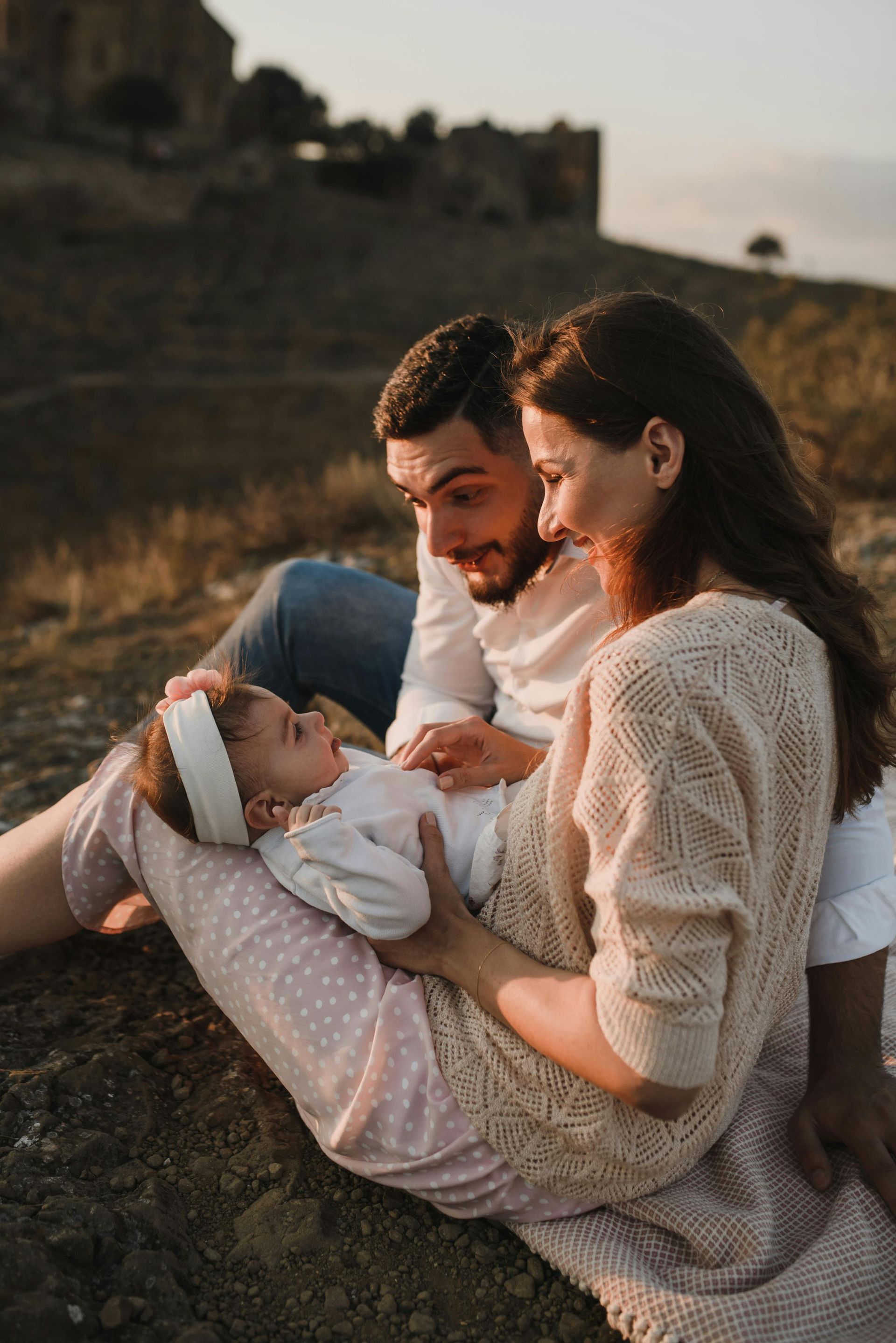 A family of three, parents looking lovingly at their baby. They're sitting outdoors, the parents smiling in the golden light of the setting sun.