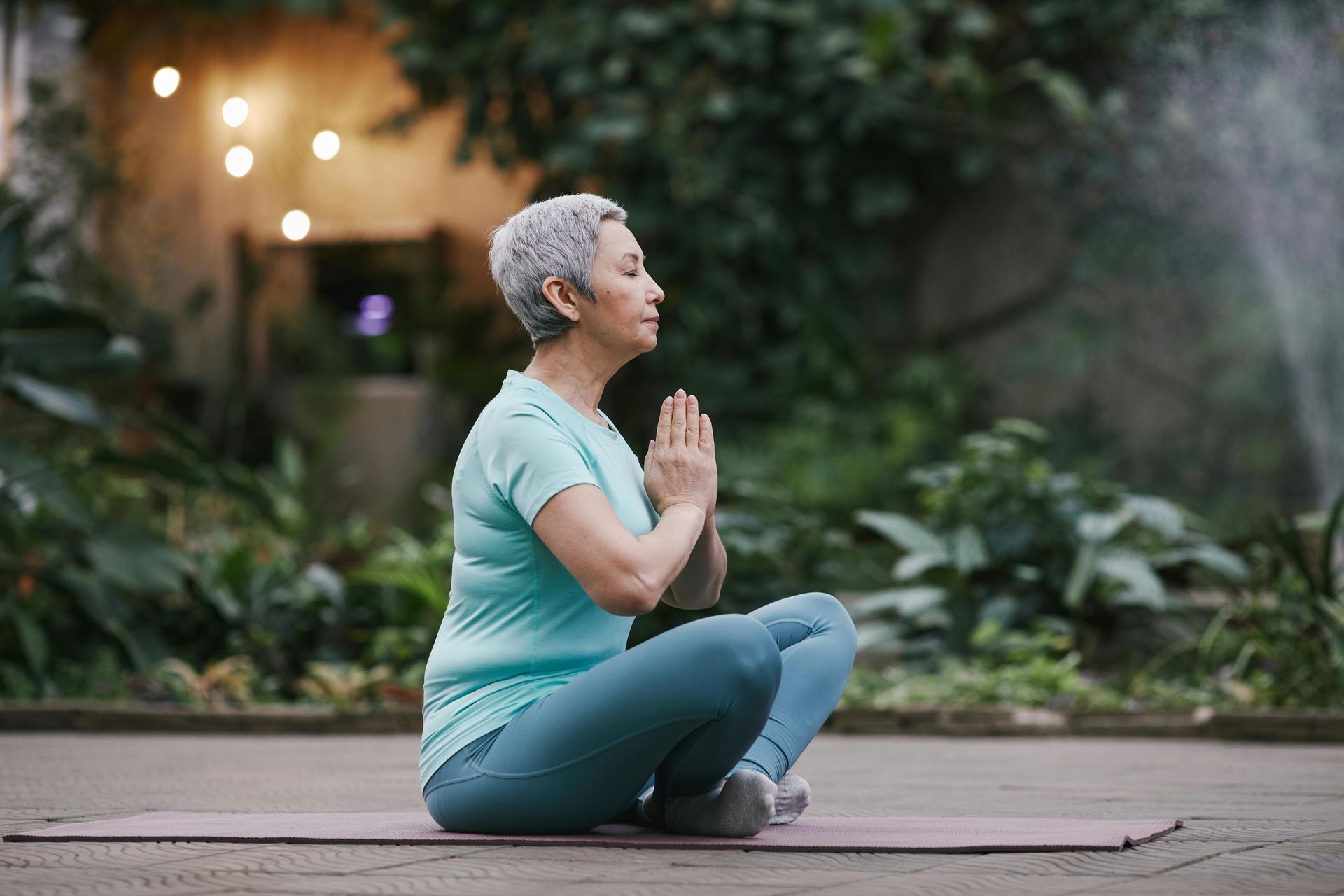 An elderly woman is sitting on a yoga mat with her hands folded in prayer.