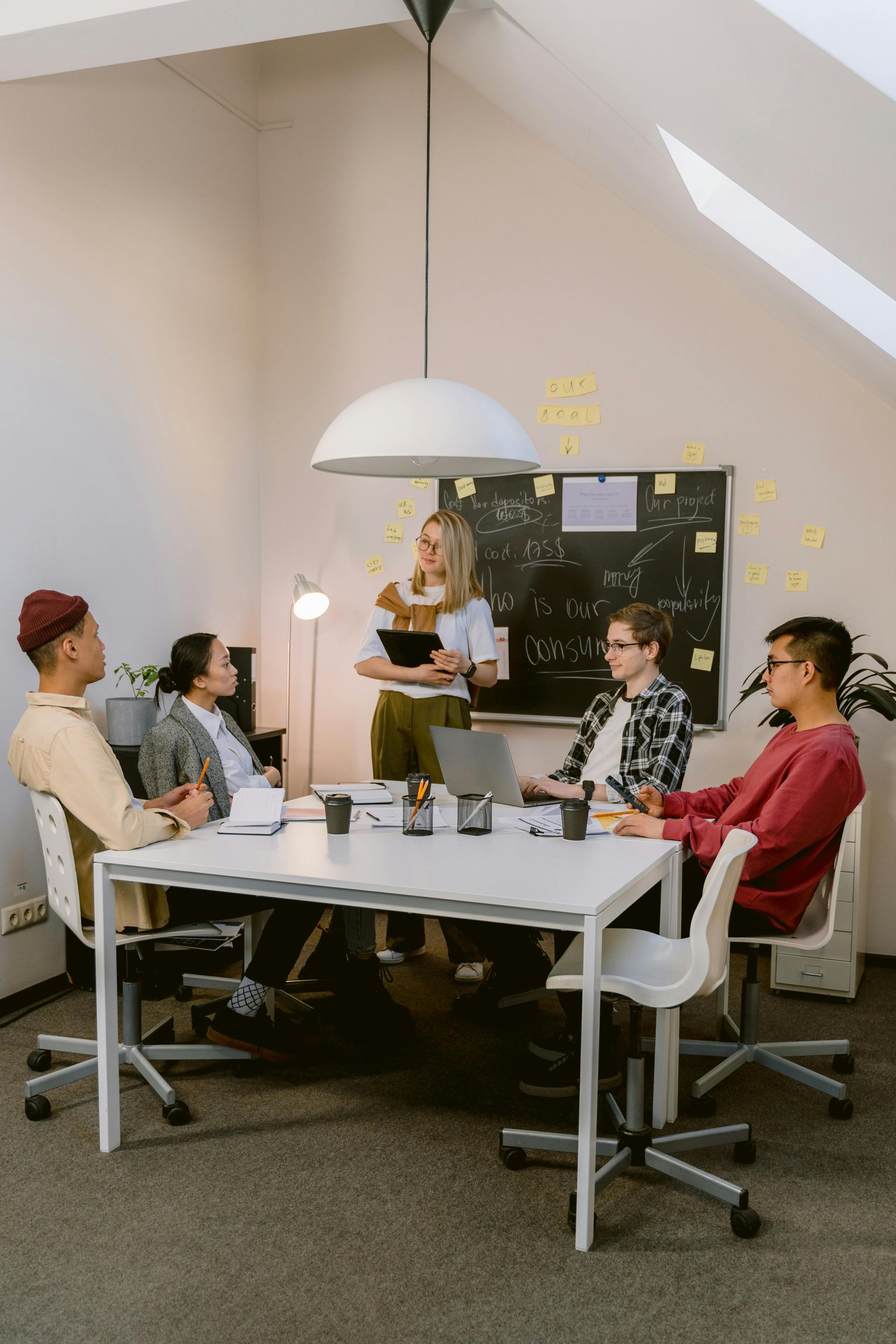 A diverse team in a bright office meeting around a white table, a woman presents a plan.