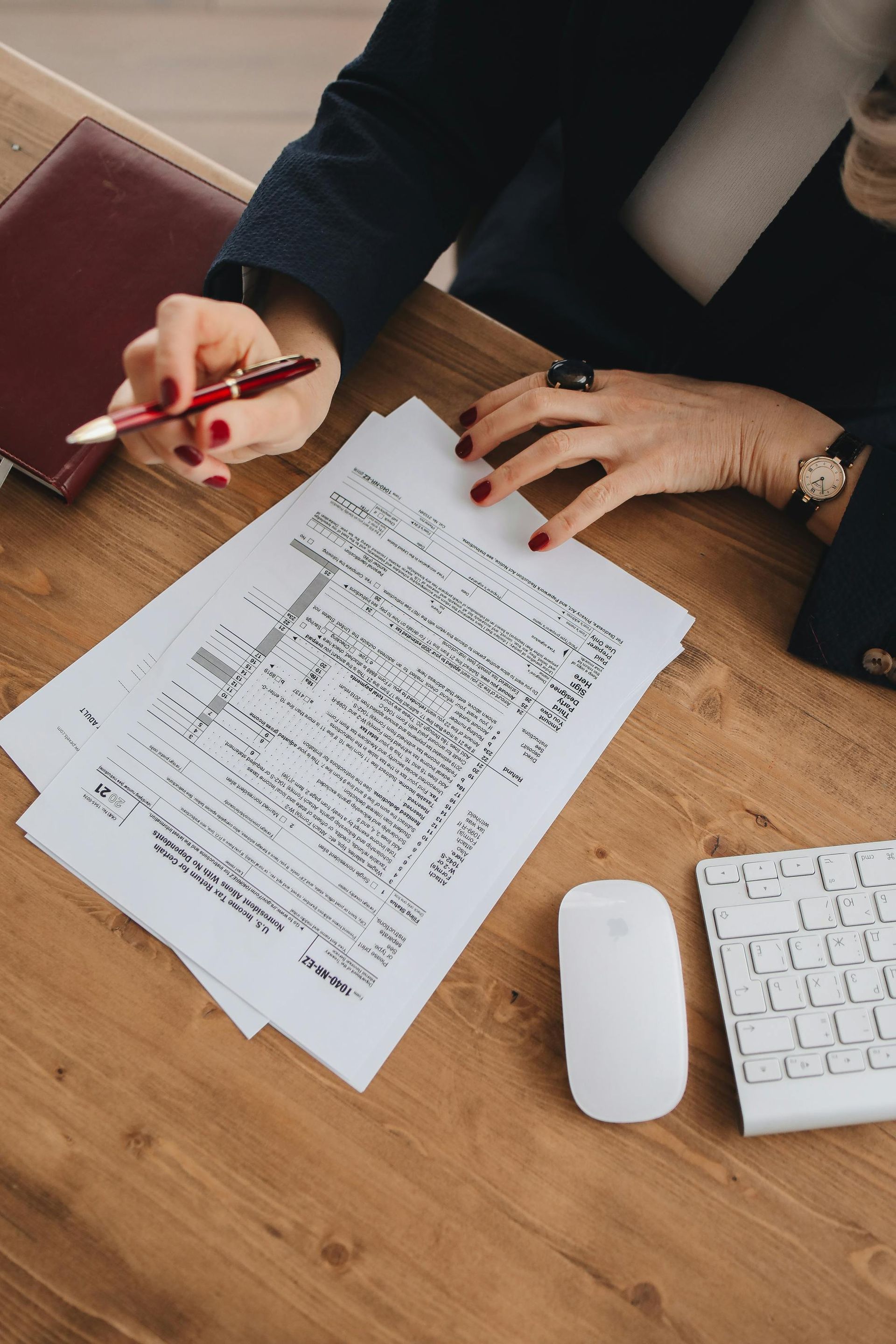 A person with red nail polish reviews documents at a desk, holding a red pen. A red notebook, white mouse, and keyboard are also on the wooden surface.