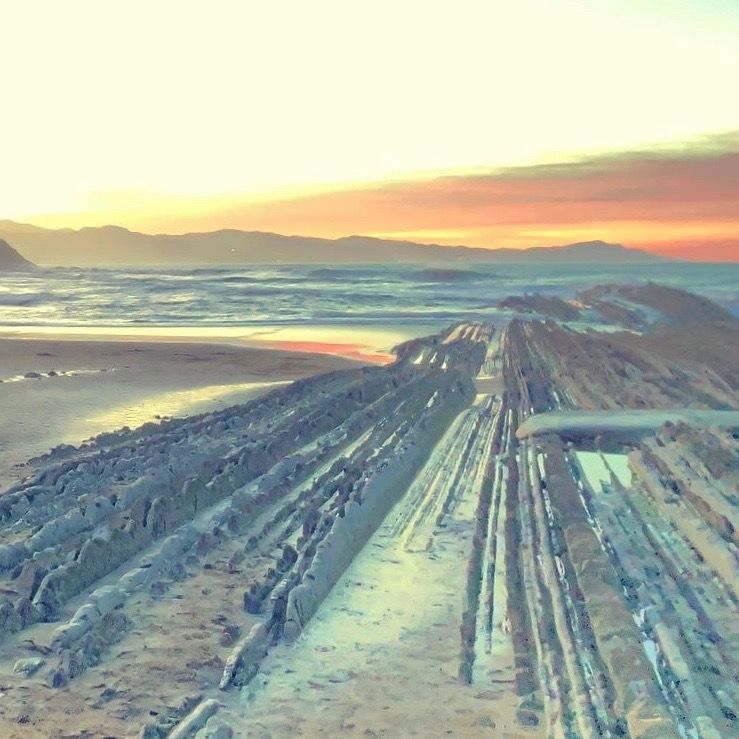 Coastal rock formations at sunset, reflecting golden light; ocean in background, mountains in distance.