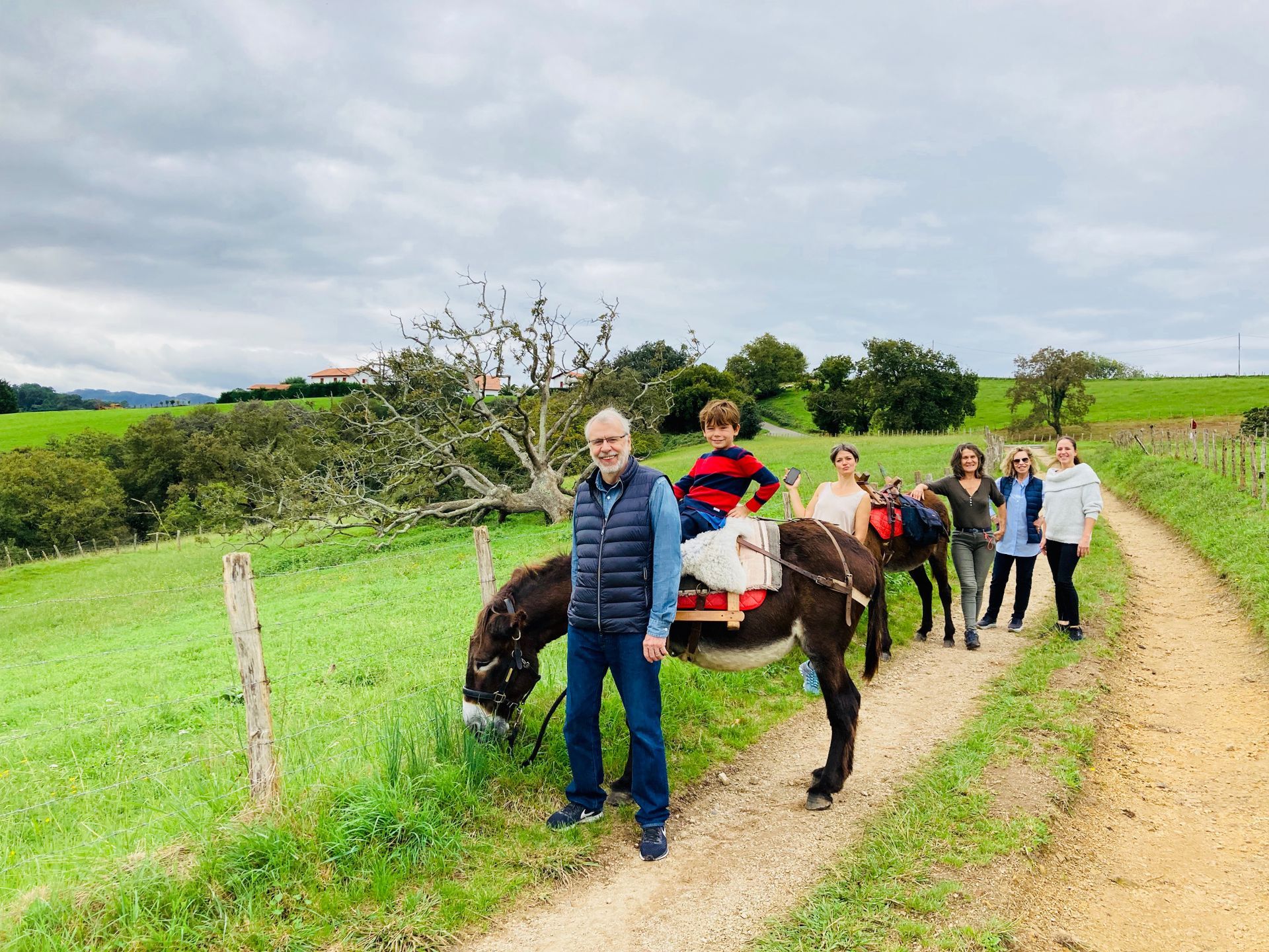 Family poses with donkeys on a grassy path. A boy rides one; adults stand nearby. Cloudy sky.