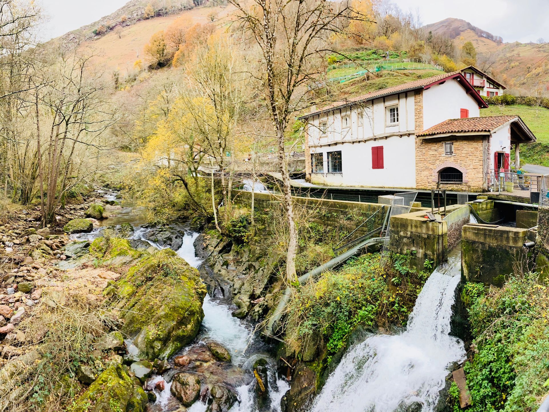 Water cascades down rocks towards a traditional building with white walls, red shutters, and a tiled roof.