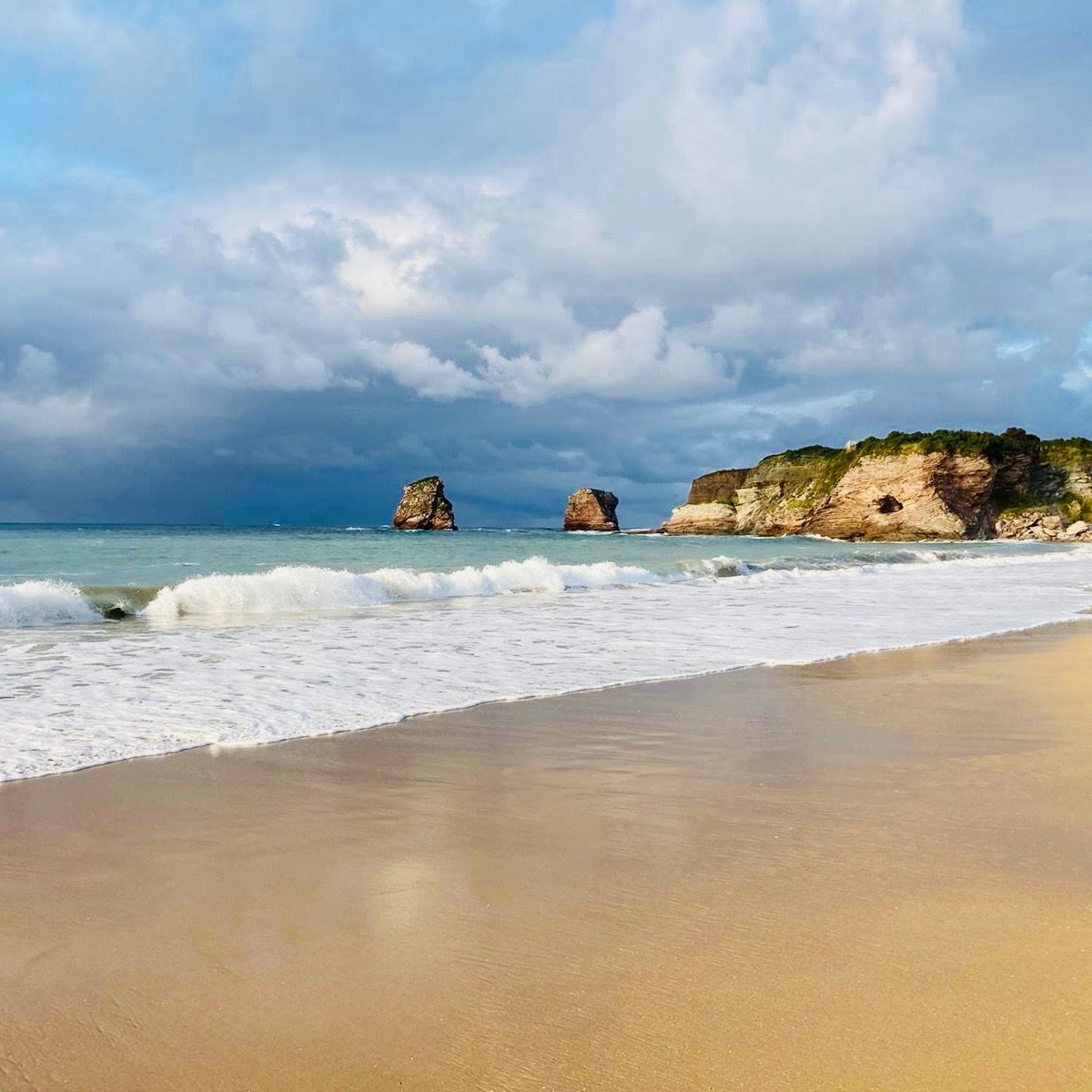 Sandy beach with waves, rocky cliffs, and cloudy sky.
