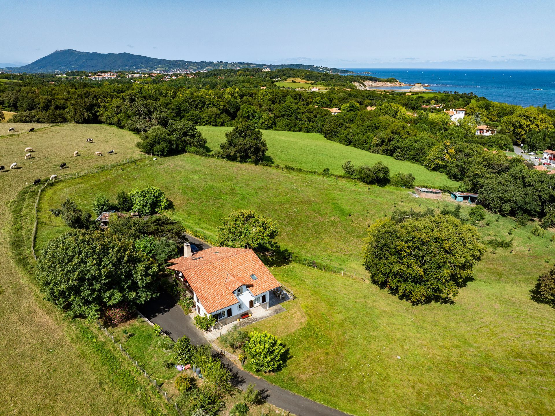 A small house with a red-tiled roof sits in a green field near a coastline, with the sea in the background.