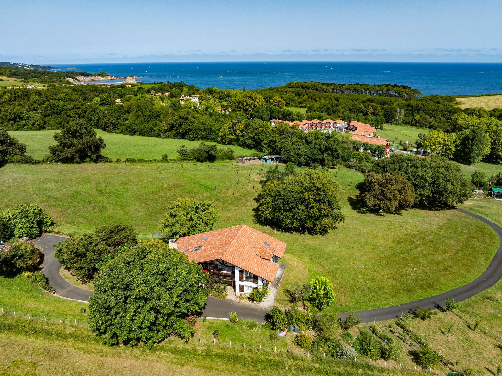 House with red tile roof, green fields, and ocean view on a sunny day.