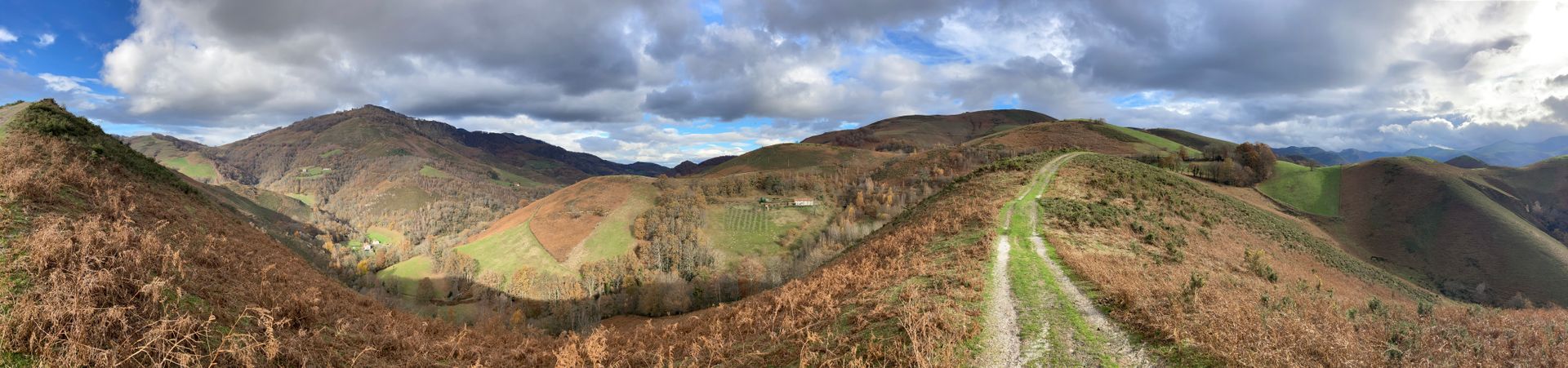 Panoramic view of rolling hills covered in brown and green vegetation under a cloudy sky. A path leads through the center.