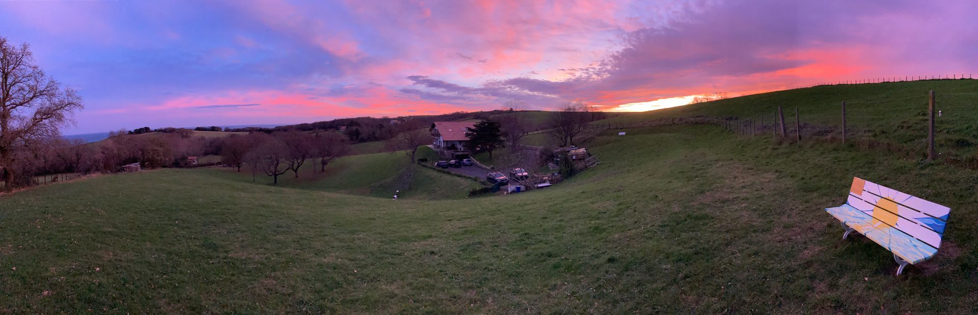 Sunset over a grassy field with trees, a building, and a bench. The sky is vibrant with pink and orange hues.