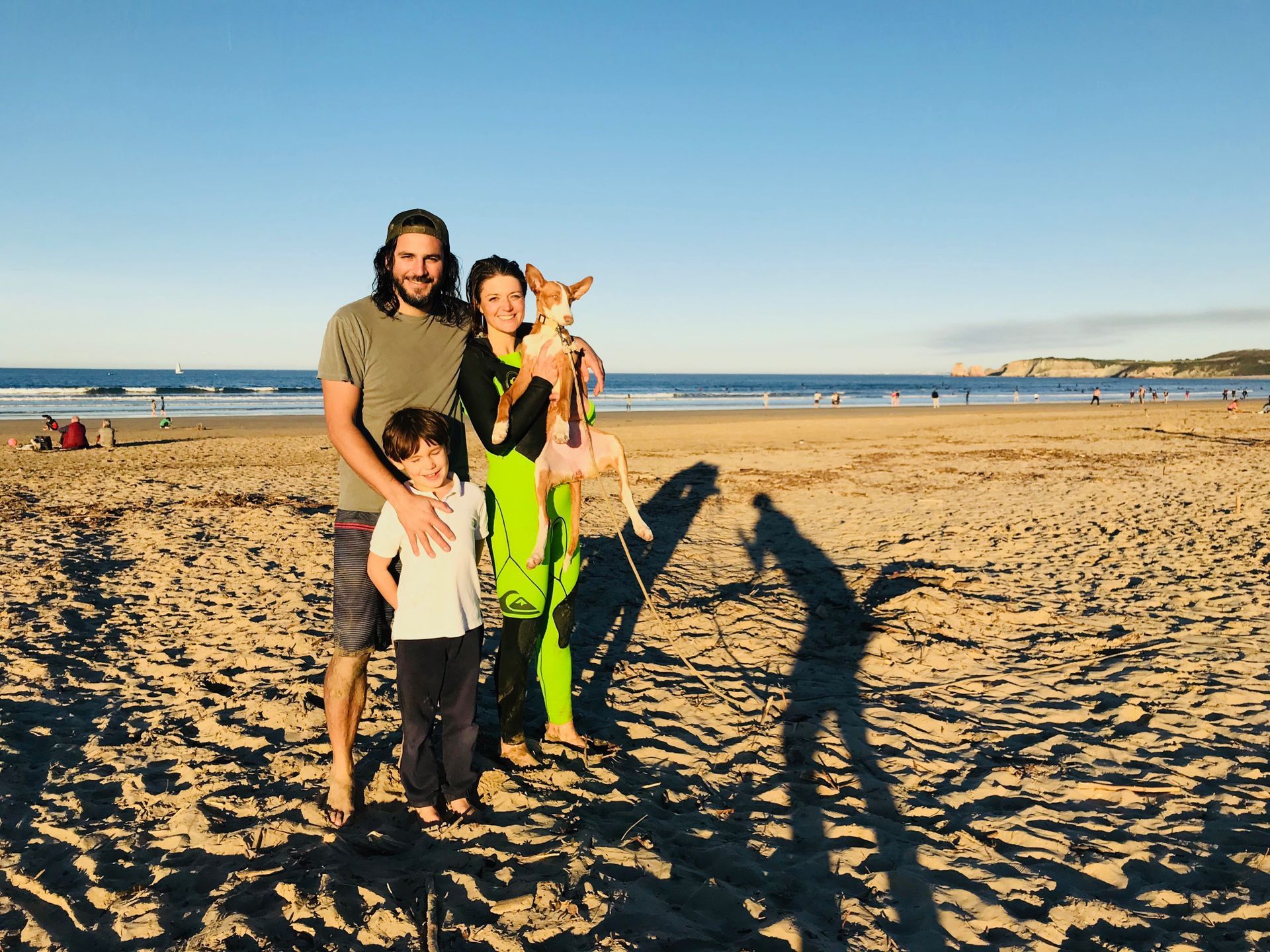 Family on beach: man, woman holding dog, child. Sunny day, shadows, ocean background.