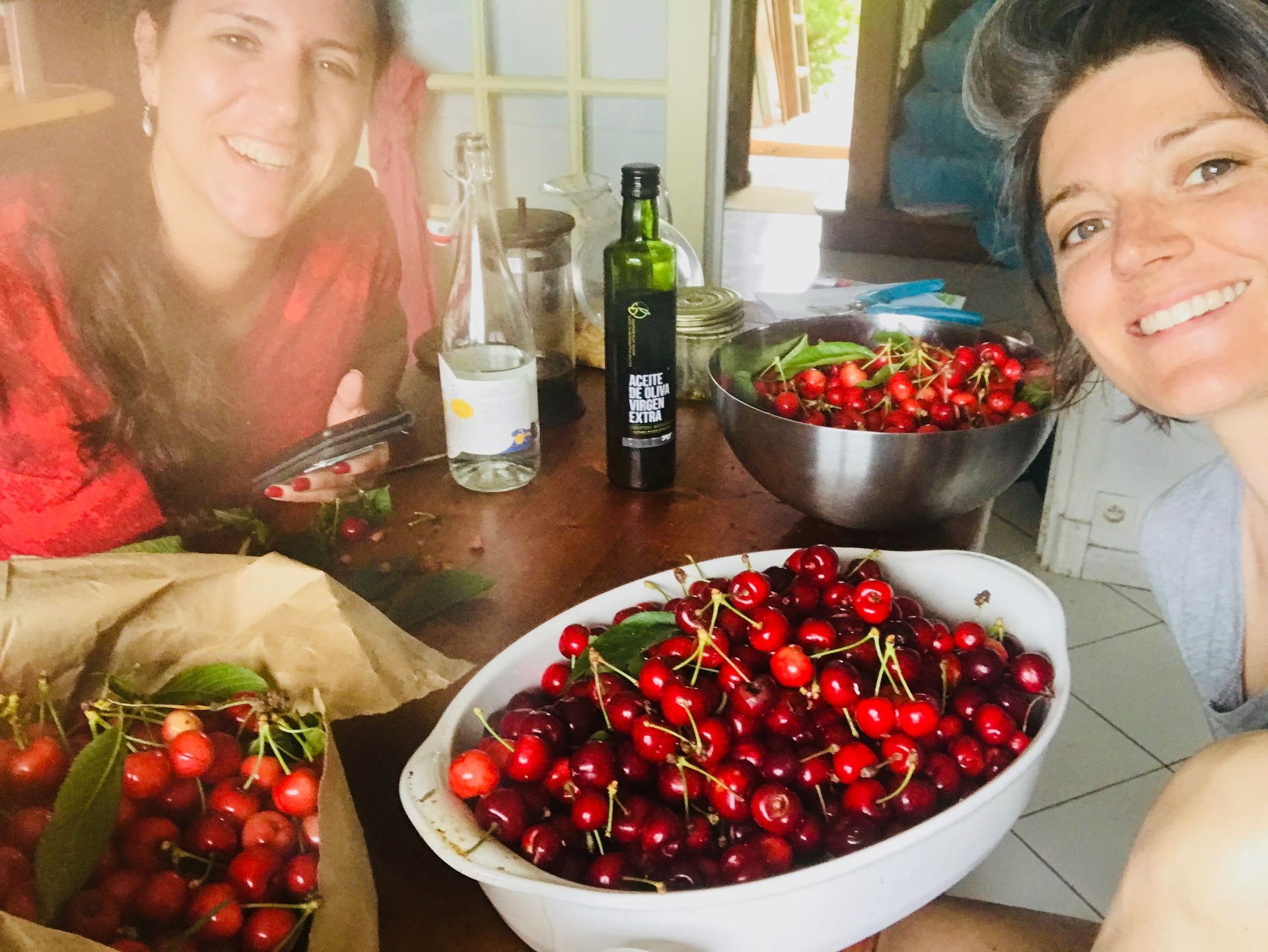 Two women smiling, surrounded by bowls and boxes of red cherries on a wooden table.