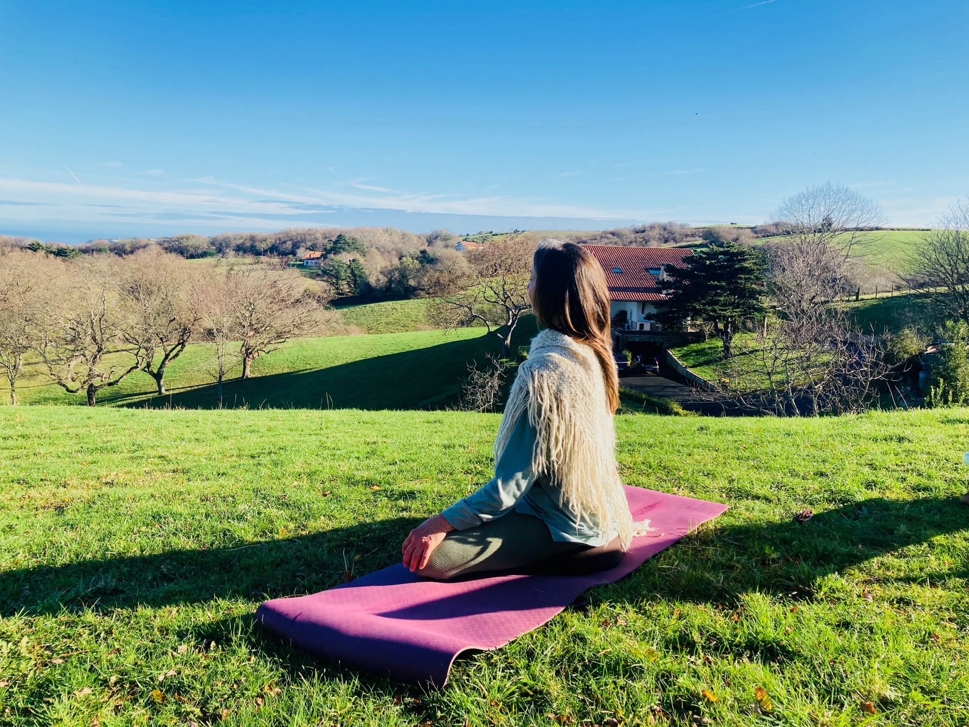 Woman meditating outdoors on a purple mat in a sunny field, facing a landscape with trees.
