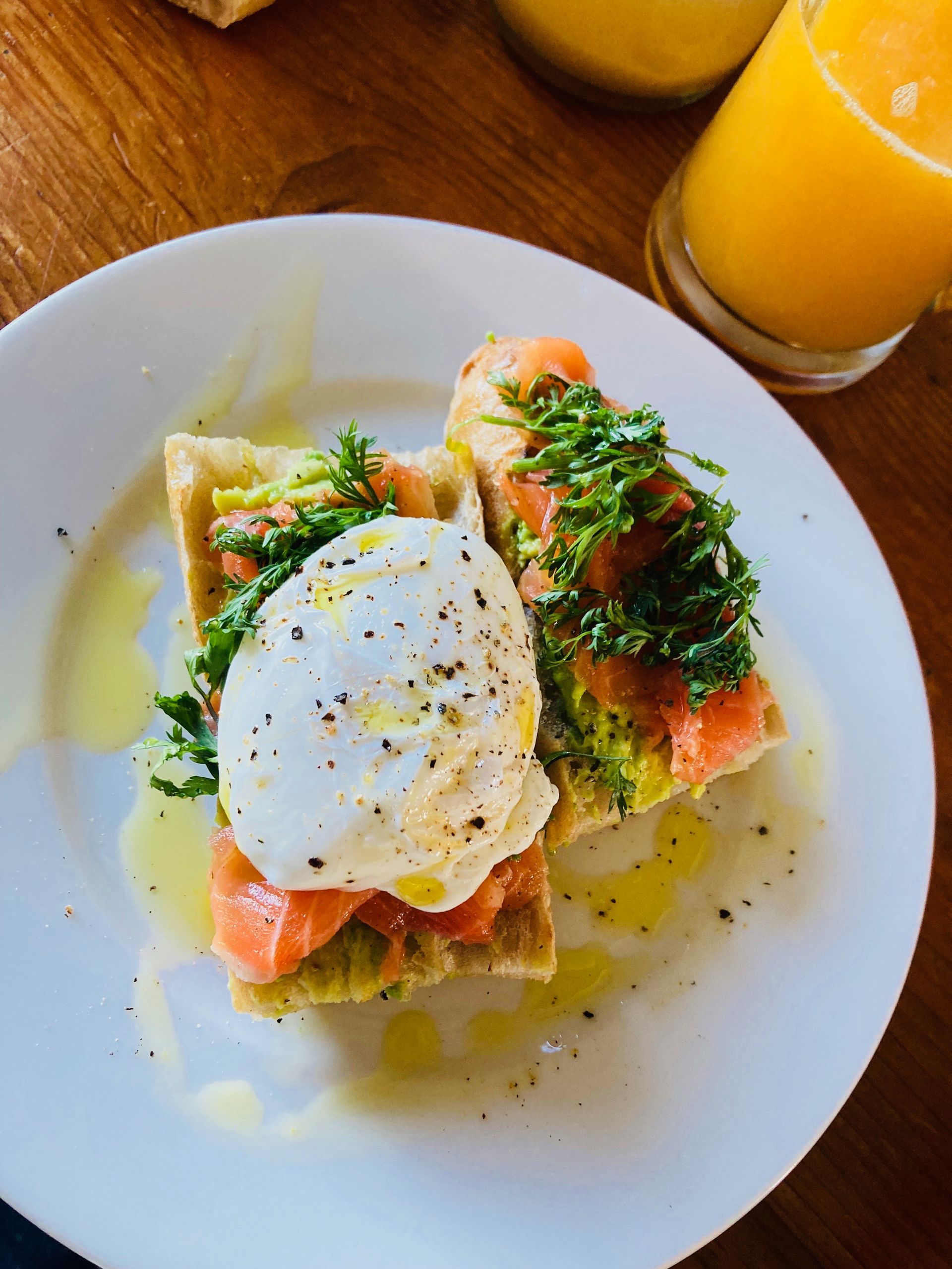 Toast topped with salmon, avocado, poached egg, and herbs, drizzled with oil, next to orange juice.